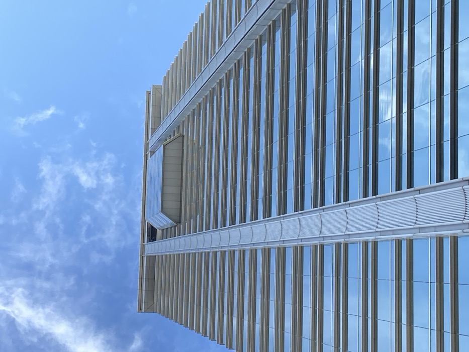 The low-angle shot showcases a modern high-rise building against a blue sky with scattered clouds. The building's facade features vertical lines of glass and metal, with a cantilevered section extending outward. Horizontal architectural elements bisect the verticality, adding depth and dimension. Reflections of the sky are visible in the glass panels, mirroring the surrounding environment.