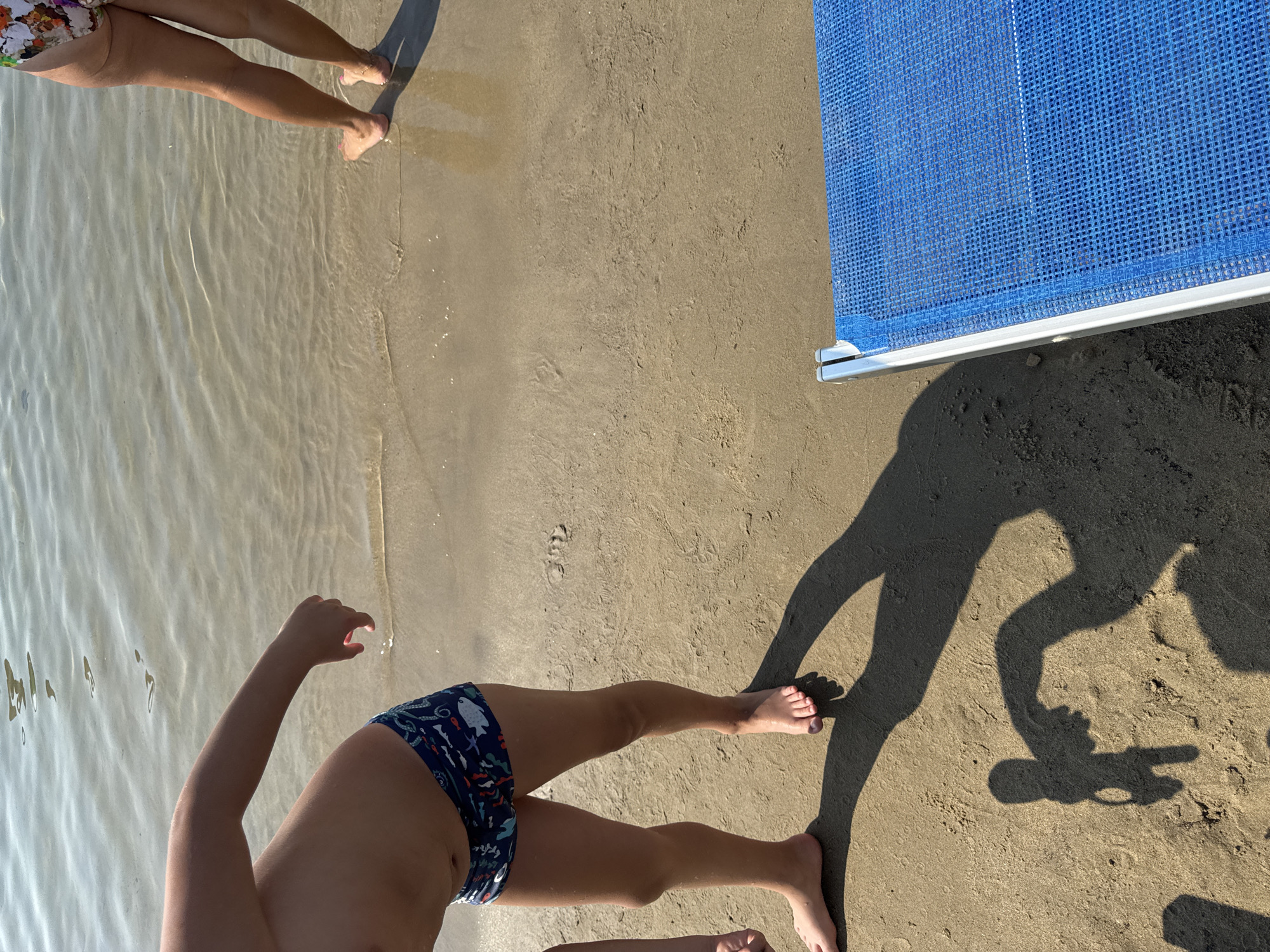 The image depicts a close-up view of a sandy beach with shallow water. In the foreground, the legs of a child wearing blue swim trunks with an underwater pattern are visible, standing with their feet in the water. A long shadow of the child is cast on the sand, extending to the left. To the right, the legs of another person, presumably an adult, are seen from the knees down, wearing a floral swimsuit, with their feet also in the water. A blue mesh beach chair is partially visible in the lower right corner, on the sand. The water is clear and calm, with reflections of sunlight on its surface. The sand is light brown and appears wet near the water's edge. There are no identifiable landmarks that place this scene specifically in Sanremo, Italy.
