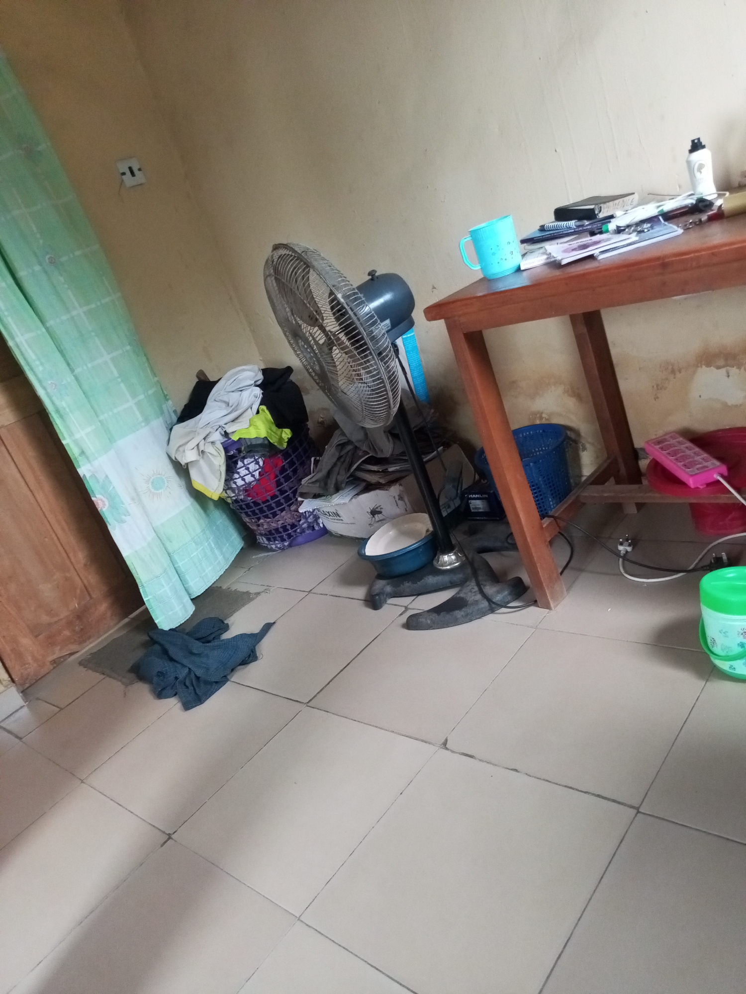 The image depicts an indoor scene, likely a room in a residential setting in Ihalav, Nigeria. The room is sparsely furnished, with a wooden table dominating the center. On the table are several books and papers, a blue plastic mug, and a white bottle. To the left of the table, a standing fan is positioned, its metal blades visible. Near the fan, a laundry basket overflowing with clothes is situated on the floor, next to a light green curtain hanging from a rod. A pile of dark blue cloth lies on the tiled floor in the foreground, partially covering a doormat. To the right of the table, a blue plastic basket and a pink tray with what appear to be ice cube molds are visible. A red bucket is also present, along with a green plastic container in the lower right corner. The walls are a light tan color, and the floor is covered with beige tiles. An electrical outlet is visible on the wall to the left. The overall impression is one of a functional, lived-in space.