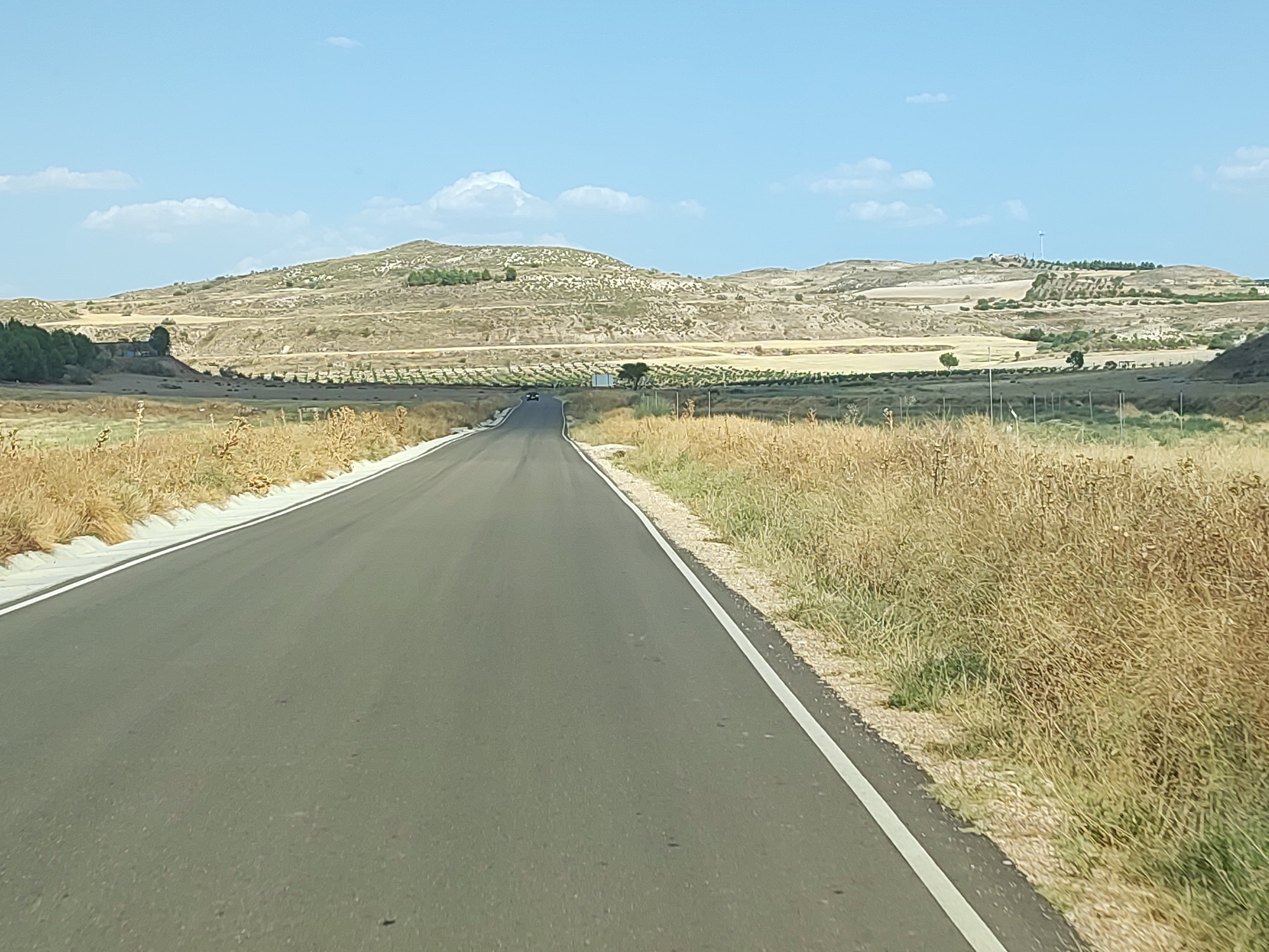 A paved road with a white line on the right edge stretches into the distance under a clear blue sky with scattered white clouds. The road is bordered by dry, golden grass on both sides. In the background, rolling hills with sparse vegetation and some trees are visible. A few structures and a wind turbine can be seen on the crest of one of the hills. The scene appears to be an agricultural or rural area in Illana, Spain, under bright daylight.
