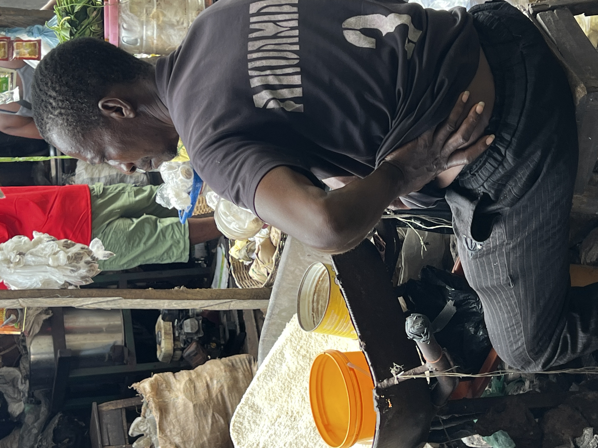 A man is crouching in an informal market setting in Nkpor, Nigeria. He is dark-skinned, wearing a dark t-shirt with light-colored text, and pinstripe pants. His left hand is placed on his right chest. He is looking down at the immediate vicinity. Around him are shelves, containers, machinery, plastic bags, and assorted objects commonly found in a marketplace. The environment is cluttered and appears to be indoors.