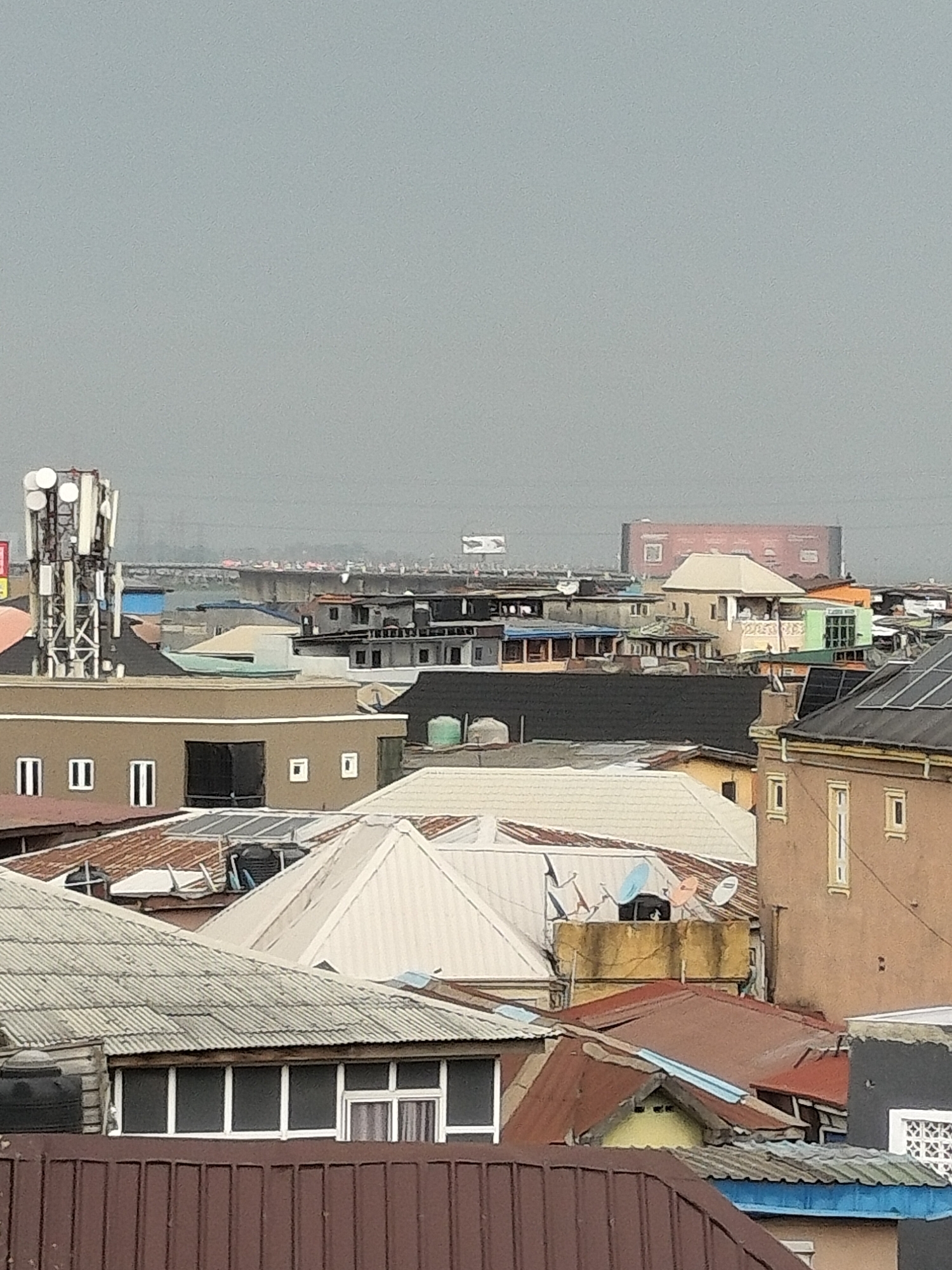 A daytime, high-angle view of a densely packed urban landscape, likely in Lagos, Nigeria. The scene is dominated by a variety of rooftops, characterized by different colors, materials (corrugated metal, tile), and architectural styles. A bridge spans the horizon in the distance, partially obscured by haze. Communication towers stand prominently on one of the buildings. Several satellite dishes are visible on rooftops. The sky is overcast, contributing to a muted color palette. The overall impression is that of a crowded and active city.