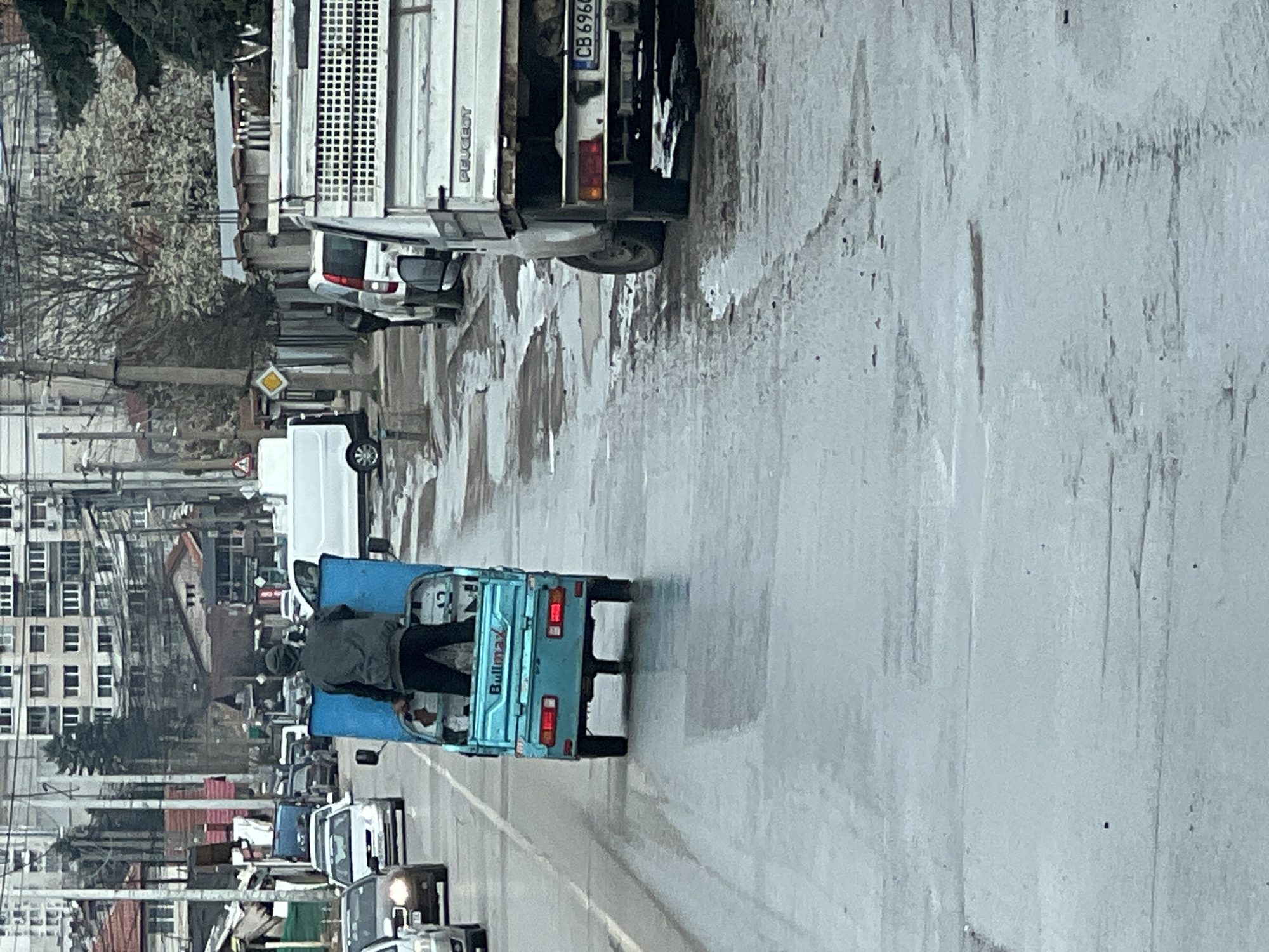 A partially obscured, street-level photograph depicts a street scene in Sofia, Bulgaria. Traffic moves on a wet road; a light blue Bullmax three-wheeled vehicle occupies the center lane, carrying two individuals on the back. The vehicle is traveling in the direction of the camera. The rear of a white truck with the license plate "CB 6966" is visible in the upper-right. Other cars are further down the road, some in motion and others parked. On the left side of the frame, there's a view of buildings with trees and streetlights. A yellow diamond-shaped road sign is visible on the left side of the street. The overall weather conditions seem overcast.