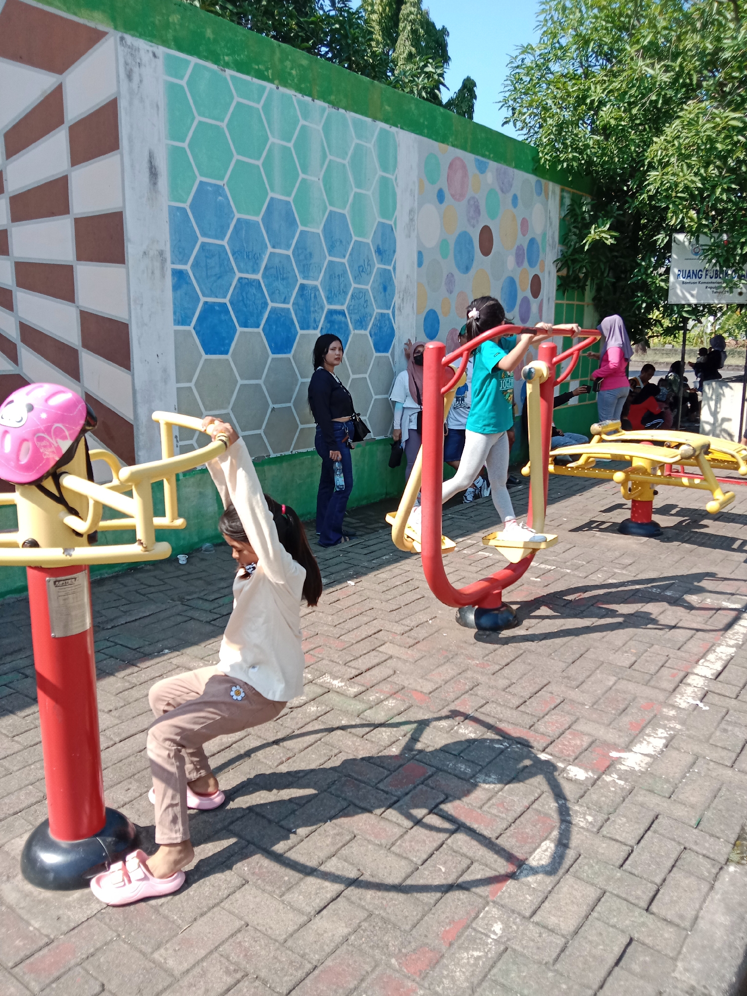 The image shows an outdoor park with exercise equipment. In the foreground, a young girl in a light-colored shirt and brown pants is using a yellow and red piece of equipment, holding onto the handles above her head. She is wearing pink sandals. To her right, another young girl in a turquoise shirt and light gray leggings is on a similar piece of red and yellow exercise equipment. Behind them, several other people are visible, some standing and others sitting. The background features a colorful mural on a wall, with geometric patterns and circular designs. Trees and a sign that reads "RUANG PUBLIK KREATIF" are also present, indicating this is likely a public recreational space. The ground is paved with interlocking bricks. The overall scene suggests a sunny day in a community park.