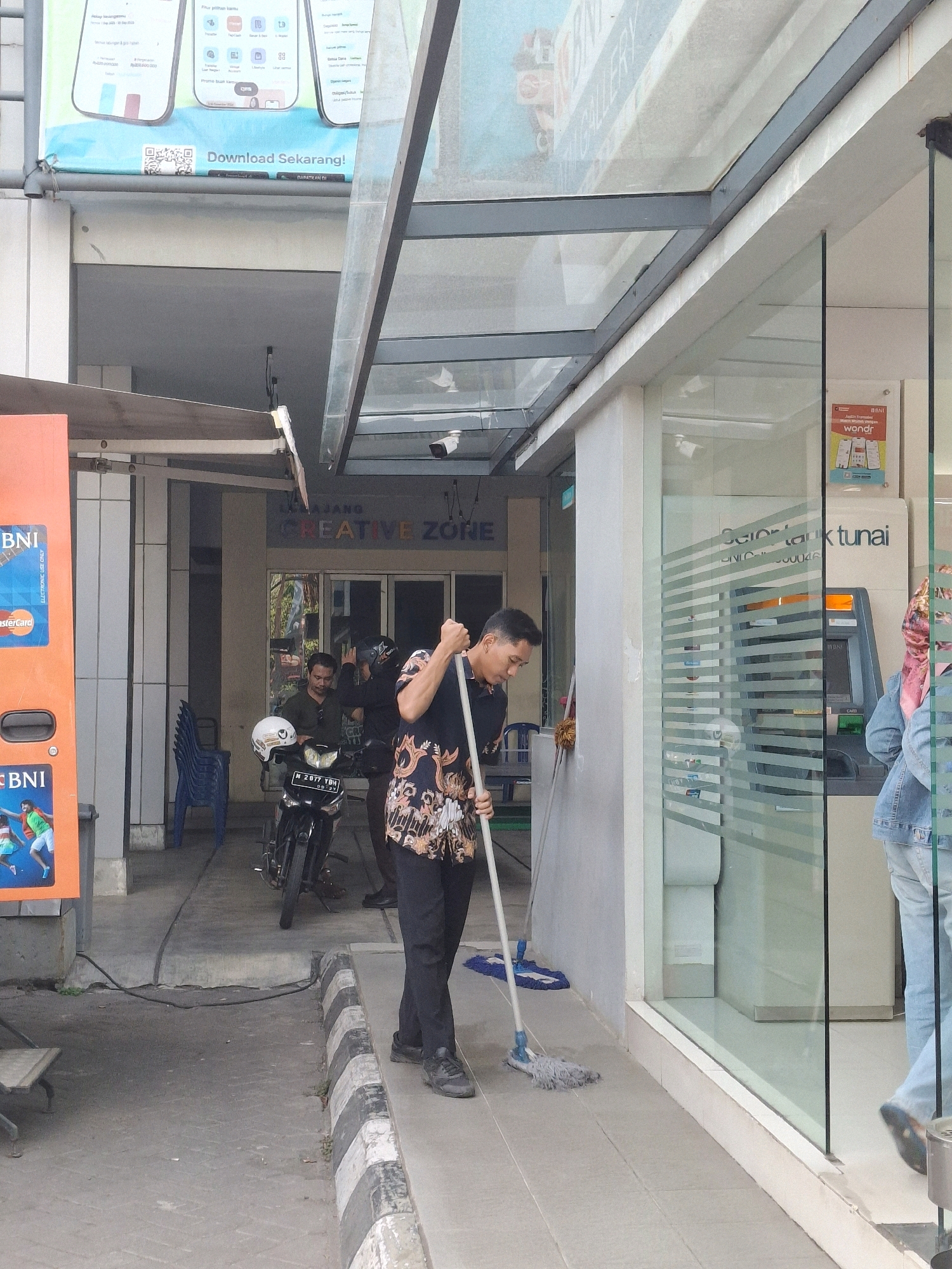 In Lumajang, Indonesia, in front of a BNI bank and ATM, a man in a black shirt with a patterned design and black pants is mopping the concrete floor with a gray mop. He is wearing dark sneakers. Behind him, to the left, is a motorcycle with two people on it, one wearing a dark shirt and the other a helmet and a black jacket. A blue plastic chair is visible behind them. An orange BNI ATM machine with credit card logos is on the far left. The bank's entrance is to the right, featuring a glass door and ATM machines visible through the glass, along with a woman wearing a pink headscarf and a denim jacket walking away from the ATM. The overall environment appears to be an outdoor public space, possibly a sidewalk or walkway in front of a commercial establishment. The time of day appears to be daytime.