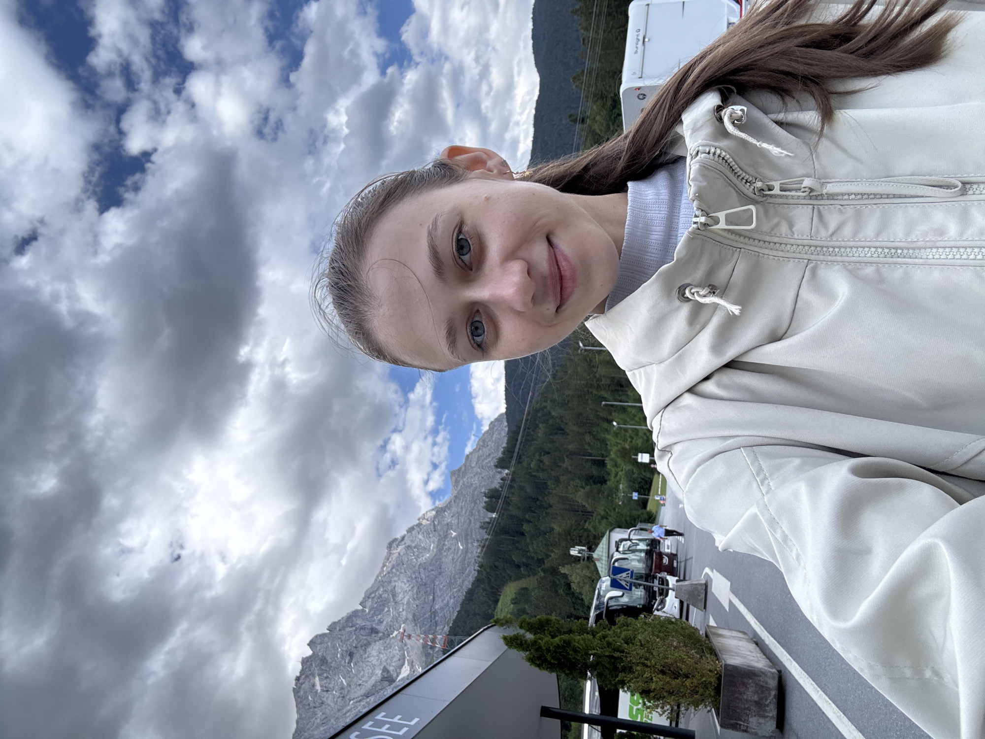 A young woman with long brown hair and blue eyes is taking a selfie outdoors. She is wearing a light-colored jacket and a grey turtleneck sweater. Behind her, there is a mountain range covered in trees, a building with the word "SEE" visible, and several buses parked. The sky is cloudy with patches of blue. The location is the Zugspitzbahn valley station in Grainau, Germany.