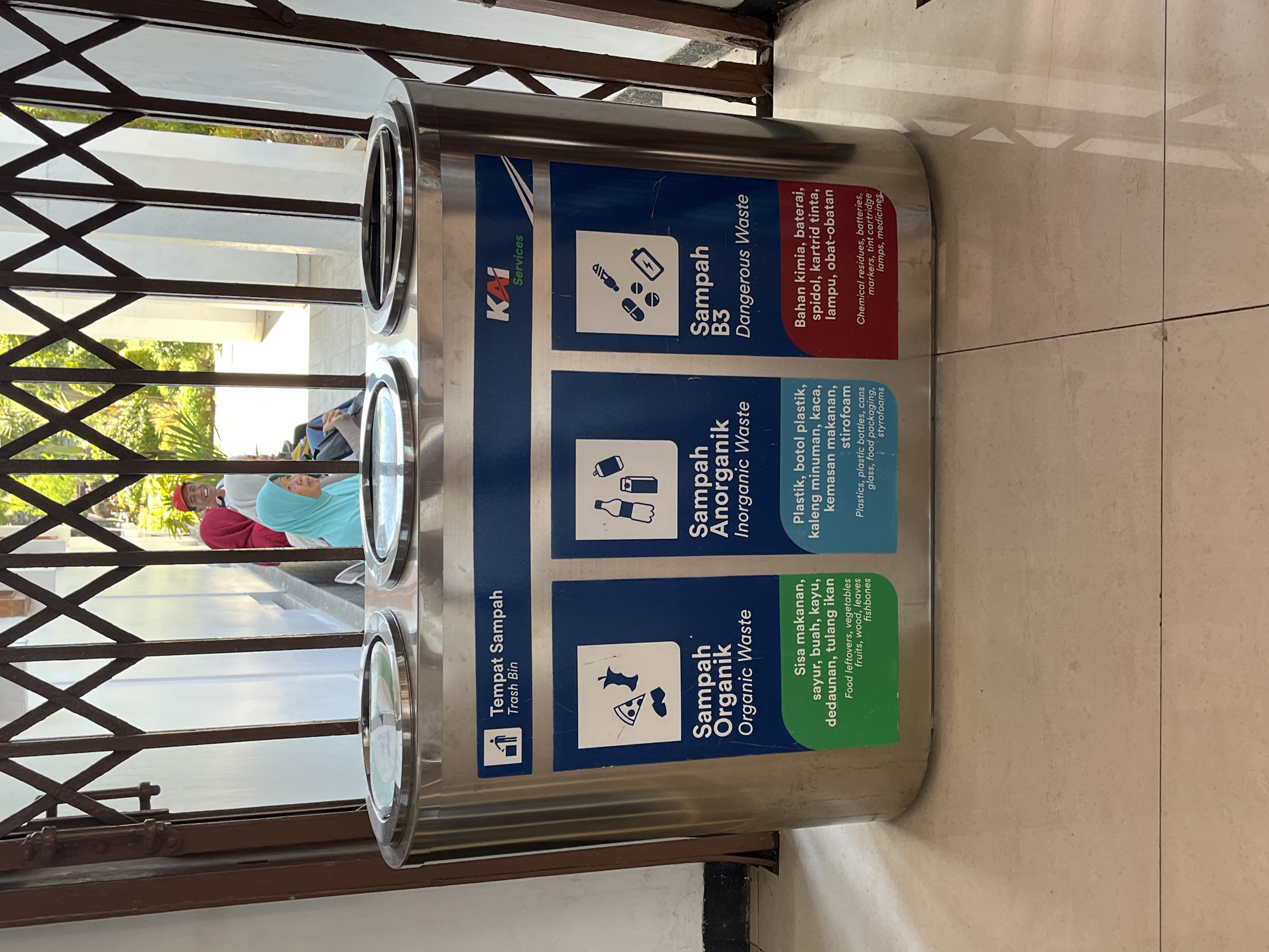 A three-compartment, stainless steel trash receptacle is centrally located in the foreground of the image. Each compartment is clearly labeled with text and icons in Indonesian and English, indicating different types of waste: "Sampah Organik" (Organic Waste), "Sampah Anorganik" (Inorganic Waste), and "Sampah B3" (Dangerous Waste). The receptacle is positioned on a tiled floor, and a metal, collapsible gate is visible behind it. Through the gate, a shaded outdoor area with white pillars and lush greenery can be seen. In the background, several individuals are visible. One person wearing a red headscarf and a red cap is smiling, while another person in a turquoise headscarf is looking to the side. The overall setting suggests an outdoor public space, possibly a transportation hub or a communal area in a place like Desa Tambakreja, Indonesia, given the visible signage and cultural context.