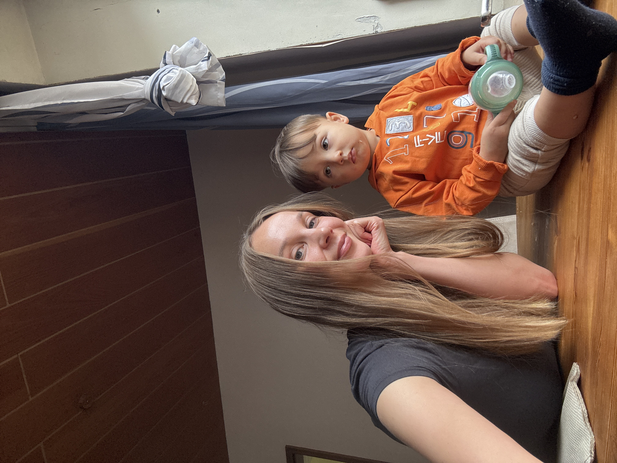 A medium shot captures a woman and a young child sitting on a wooden surface, likely a table, indoors. The woman, on the left, is looking towards the camera with a slight smile, resting her chin on her hand. She has long, blonde hair and is wearing a dark t-shirt. To her right, the child, a toddler with brown hair, is looking slightly upwards and to the left. The child is wearing an orange long-sleeved shirt with graphics and light-colored pants. The child is holding a green sippy cup. In the background, a window with curtains is visible, and the ceiling appears to be made of wood paneling. The overall lighting suggests it is daytime. The image was taken in Überherrn, Germany.