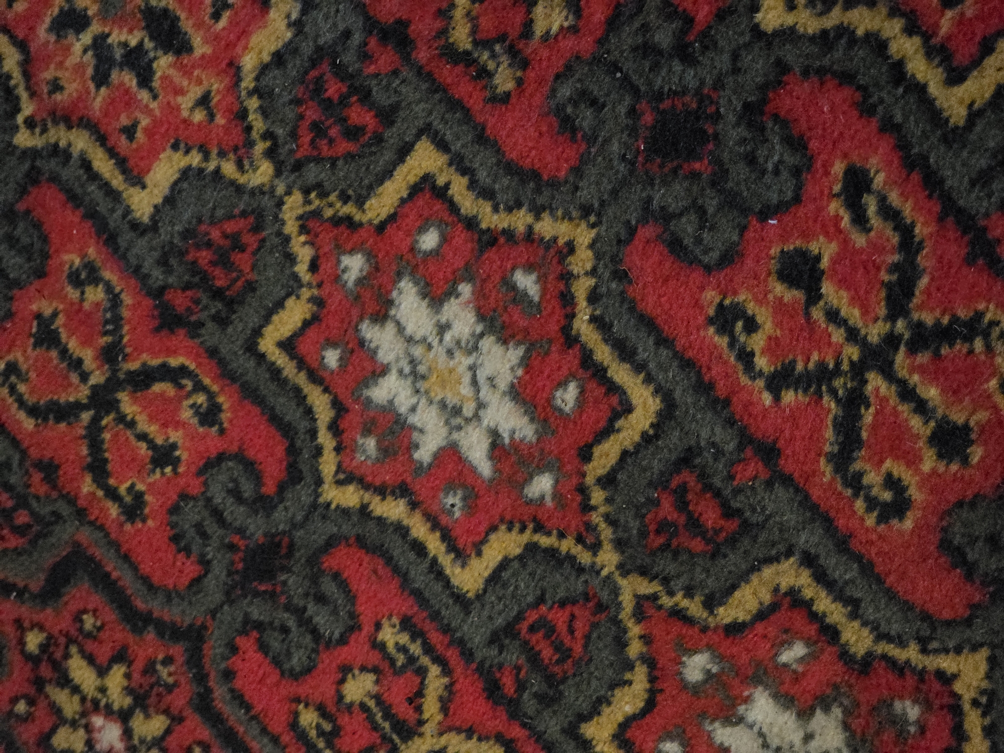 The image is a close-up, high-angle shot of a carpet with a complex geometric pattern. The dominant colors are red, black, and yellow, with some areas of white or cream. The pattern features intricate, repeating motifs that appear to be floral or star-shaped, outlined in black and yellow, and set against a red background. The texture of the carpet is visible, showing the woven fibers. The image is devoid of any context related to Petrushenko, Russia, or any discernible people, actions, or interactions. It is purely a visual study of the carpet's design and texture.