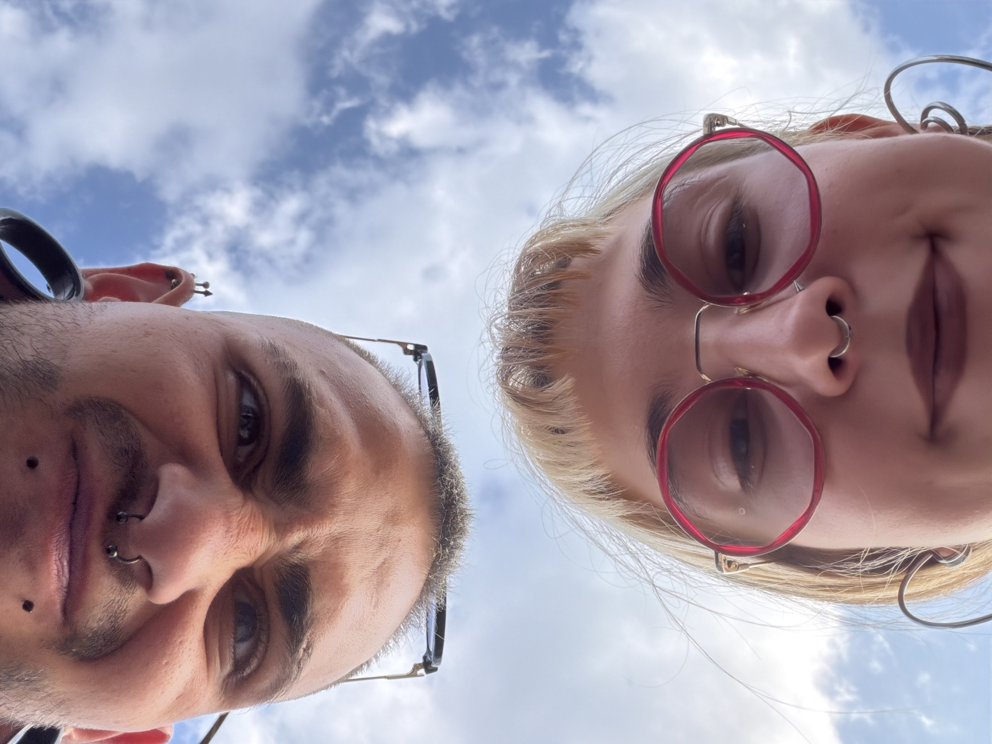 The scene takes place in Kalekovets, Bulgaria. The image is captured from a low-angle perspective, looking upwards at two individuals' faces against a bright blue sky with scattered white cumulus clouds.
In the upper part of the frame, an adult male's face is oriented upside down. He has short dark hair and stubble, with prominent eyebrows. His nose features a horseshoe-shaped septum piercing, and two small dark spots are visible above his upper lip. He wears thin, wire-frame glasses. His visible left ear (on the right side of the frame, when inverted) has multiple piercings, including one with spiked ends, and a dark, possibly plastic, ear accessory on the upper cartilage. His eyes are open, looking downwards into the camera.
Below him, an adult female's face is oriented right-side up, looking directly into the camera. She has blonde hair with bangs partially obscuring her forehead. Her nose features a small ring septum piercing. Her lips are closed and appear to have a dark color. She wears large, circular glasses with distinct red frames. Her visible left ear (on the right side of the frame) has a large, multi-loop silver-colored earring.