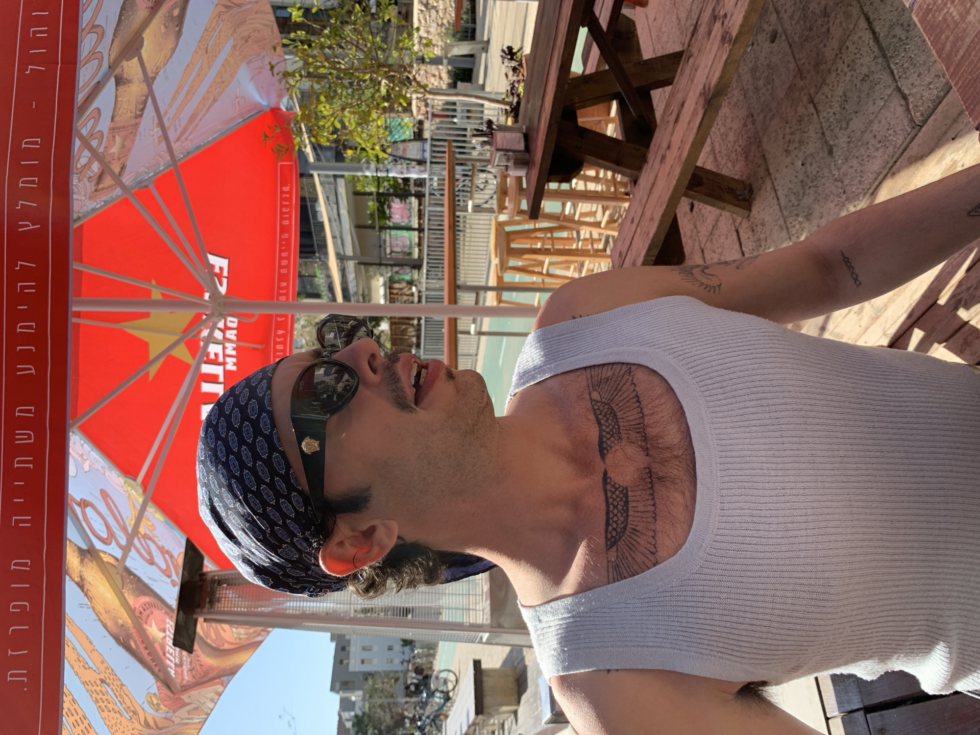 A man is sitting at a table under an umbrella, seemingly outdoors. He is wearing a white tank top, sunglasses, and a bandana. The man has tattoos on his chest and arm. A wooden picnic table sits nearby. The background suggests an urban setting, possibly Tel-Aviv, Israel, based on the text on the umbrella and the architectural style.