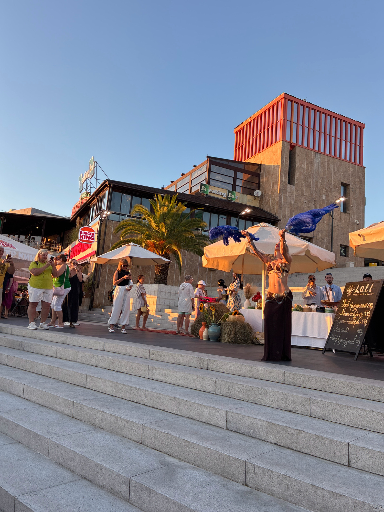 The image depicts an outdoor scene on a sunny day, likely in Sochi, Russia, given the architecture and atmosphere. In the foreground, a wide set of granite stairs leads up to a paved area where several people are gathered.
The central focus of the paved area is a woman performing what appears to be a belly dance. She is wearing a two-piece costume with intricate embellishments and a dark, flowing skirt. Her arms are raised, holding large blue feathery props. She is performing under a large, cream-colored umbrella, with a white tablecloth-covered table behind her displaying various items, possibly food or merchandise.
Several other individuals are present in the scene. To the left, a group of women are standing and observing the performance, some holding up their phones, suggesting they are taking pictures or videos. Another woman in a white top and pants stands closer to the performance, holding a drink and a phone. Further to the right, a man and a woman are engaged in conversation. In the background, a building with a distinct orange-colored upper section and stone cladding is visible, with a "Burger King" sign prominently displayed on a lower level. A large palm tree is also visible near the building, contributing to a resort-like ambiance.
A black chalkboard sign, with text written in what appears to be Cyrillic script, stands to the right of the performer. The sign displays what looks like a menu or a list of items with prices. The overall environment suggests a public gathering, possibly a festival or a market event, taking place near a commercial area. The lighting indicates it is late afternoon or early evening, with the sun casting long shadows and illuminating the building's facade.