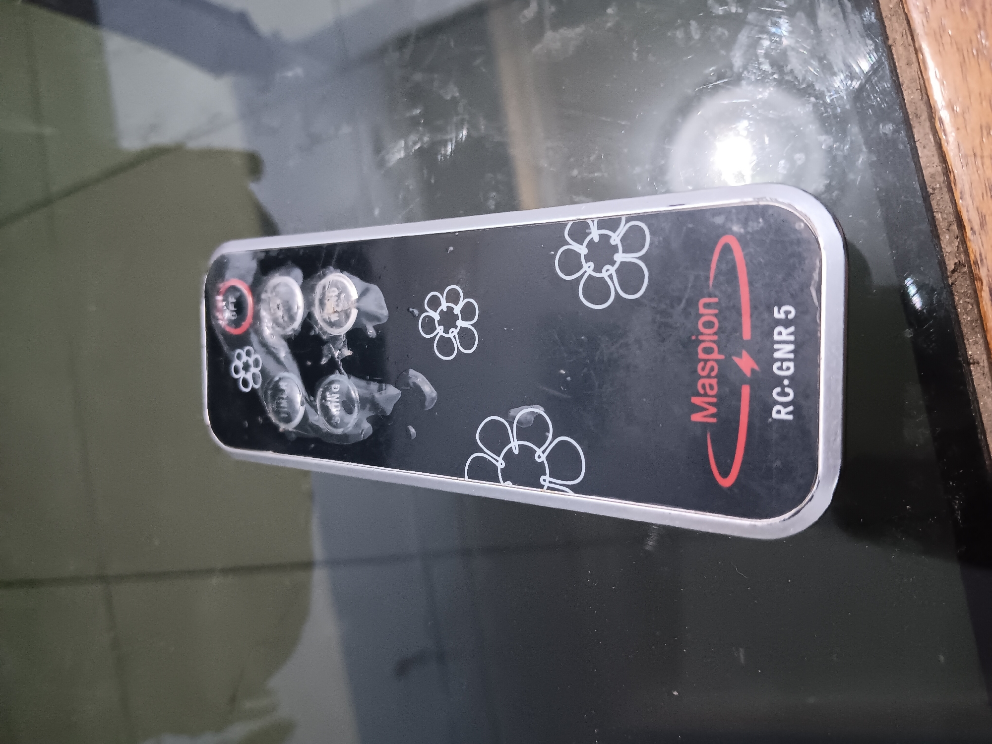 A black and silver remote control, possibly for an electronic appliance, rests on a reflective, dark surface, potentially a table. The remote features several circular buttons, some with faint text, and decorative floral outlines. A prominent red logo with the text "Maspion" and "RC-GNR 5" is visible on the lower part of the remote. The surface it's on appears somewhat dirty and scratched, suggesting everyday use. The lighting indicates an indoor setting. There is no information in the image to suggest a specific location in Batursari, Indonesia.