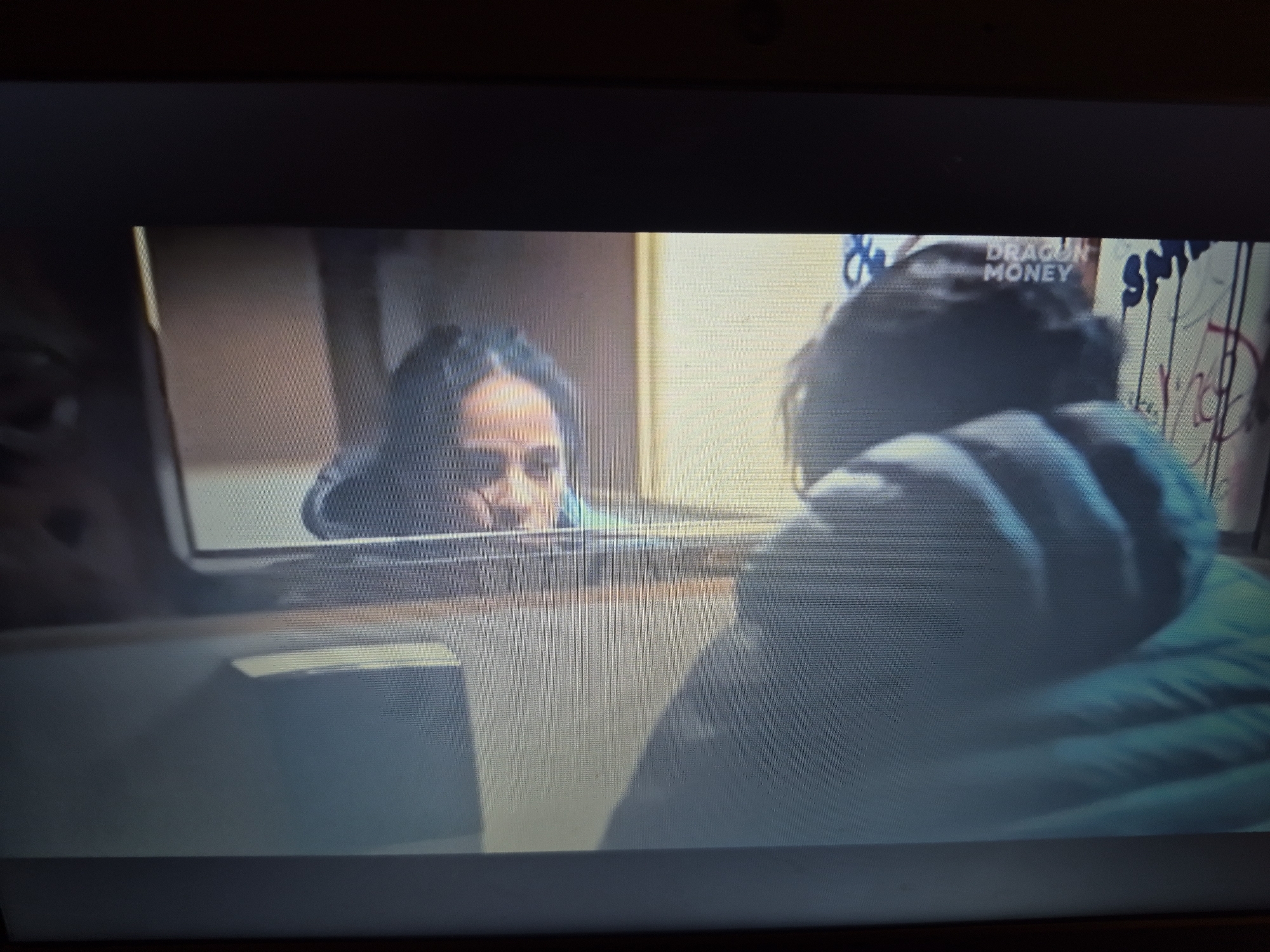 A screen capture shows a dark, slightly blurry scene of two individuals. In the foreground, on the right side of the image, a person wearing a teal puffer jacket is seen from behind, with their head turned to the left. Their dark hair is visible. In the background, a woman with dark hair is looking out from behind a translucent barrier, appearing to be in a confined space. Her face is partially obscured, but her eyes and forehead are visible. Above the woman's head, the words "DRAGON MONEY" are visible in white text. The background appears to be a wall with graffiti. The scene suggests an interaction through a window or a screen, possibly in a location within the city of Sandwell, United Kingdom, although this cannot be definitively confirmed from the image alone.