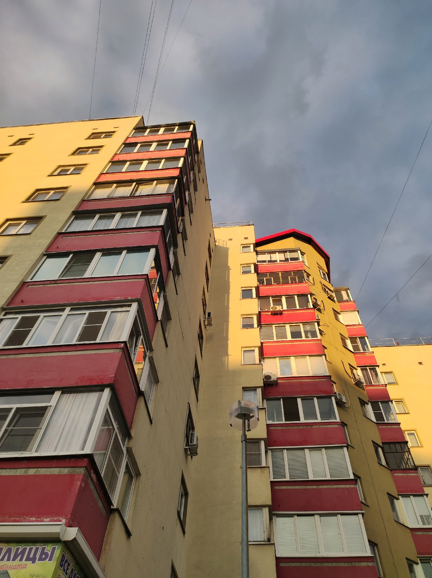 The image shows a residential building in Chaikovsky, Russia. The building is tall and appears to be a multi-story apartment complex. The facade is painted a light yellow and features red accents around the balconies and window frames. Many balconies are visible, some enclosed and others open. The windows are varied in design, with some showing blinds or curtains. The sky is overcast with dark clouds, and the sunlight is hitting the building at an angle, creating a warm glow on one side. Overhead power lines are visible against the sky. There is a street lamp in front of the building. A partial view of a sign with Russian text is visible at the bottom left of the image. There are no people or vehicles clearly visible in the scene.