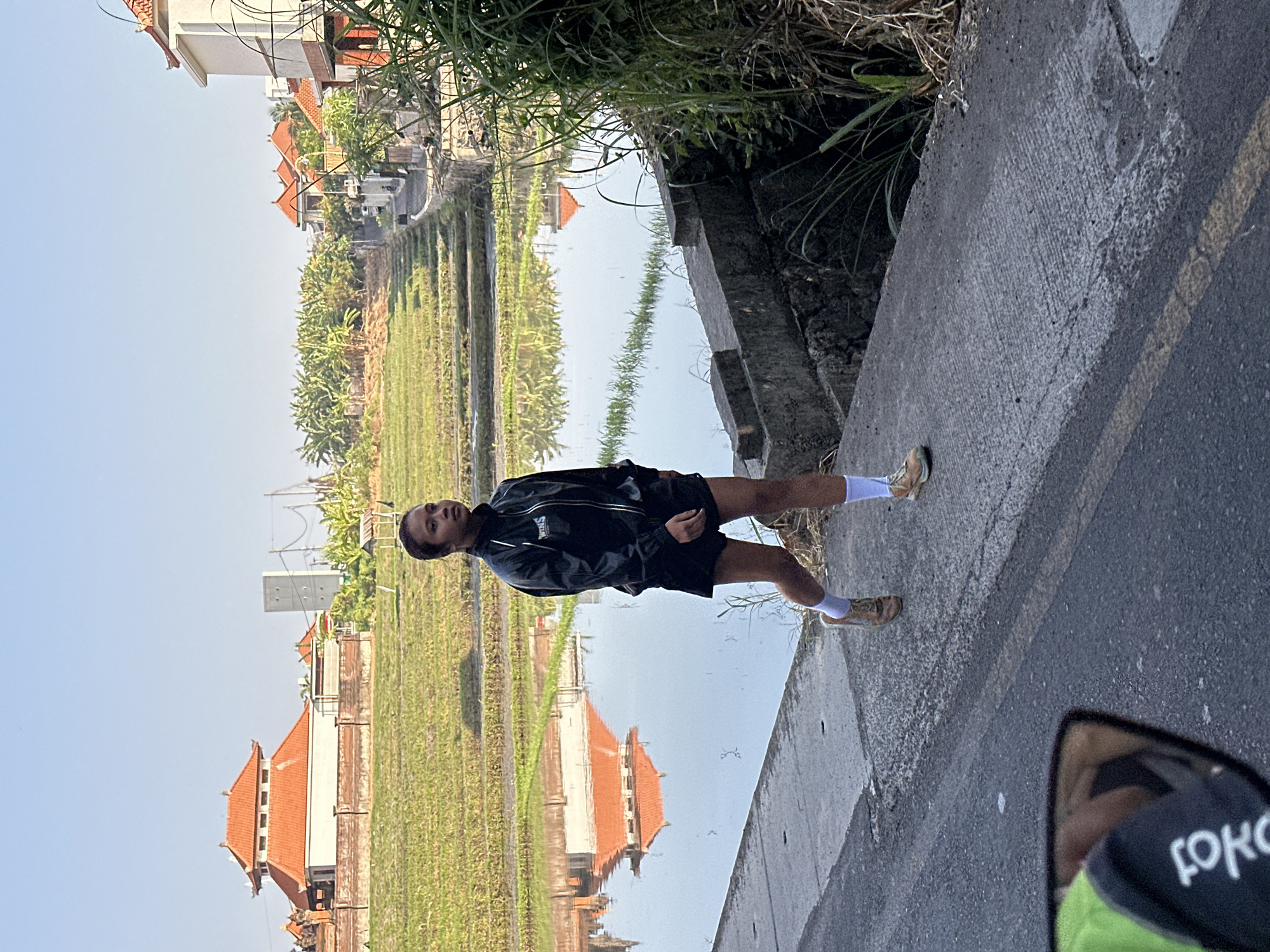 In North Kuta, Indonesia, a young woman in athletic wear walks along a paved pathway adjacent to a flooded rice paddy. She is wearing a black zip-up jacket with a white logo, black shorts, white socks, and athletic shoes. The rice paddy reflects the clear blue sky and the architecture of nearby buildings, featuring terracotta roofs. Lush green vegetation lines the far side of the paddy and grows along the embankment next to the pathway. The scene is bathed in bright sunlight, suggesting it is daytime. In the foreground, a motorcycle mirror reflects the back of someone wearing a bright green vest over black clothing. The overall environment depicts a rural or semi-rural setting within the city.