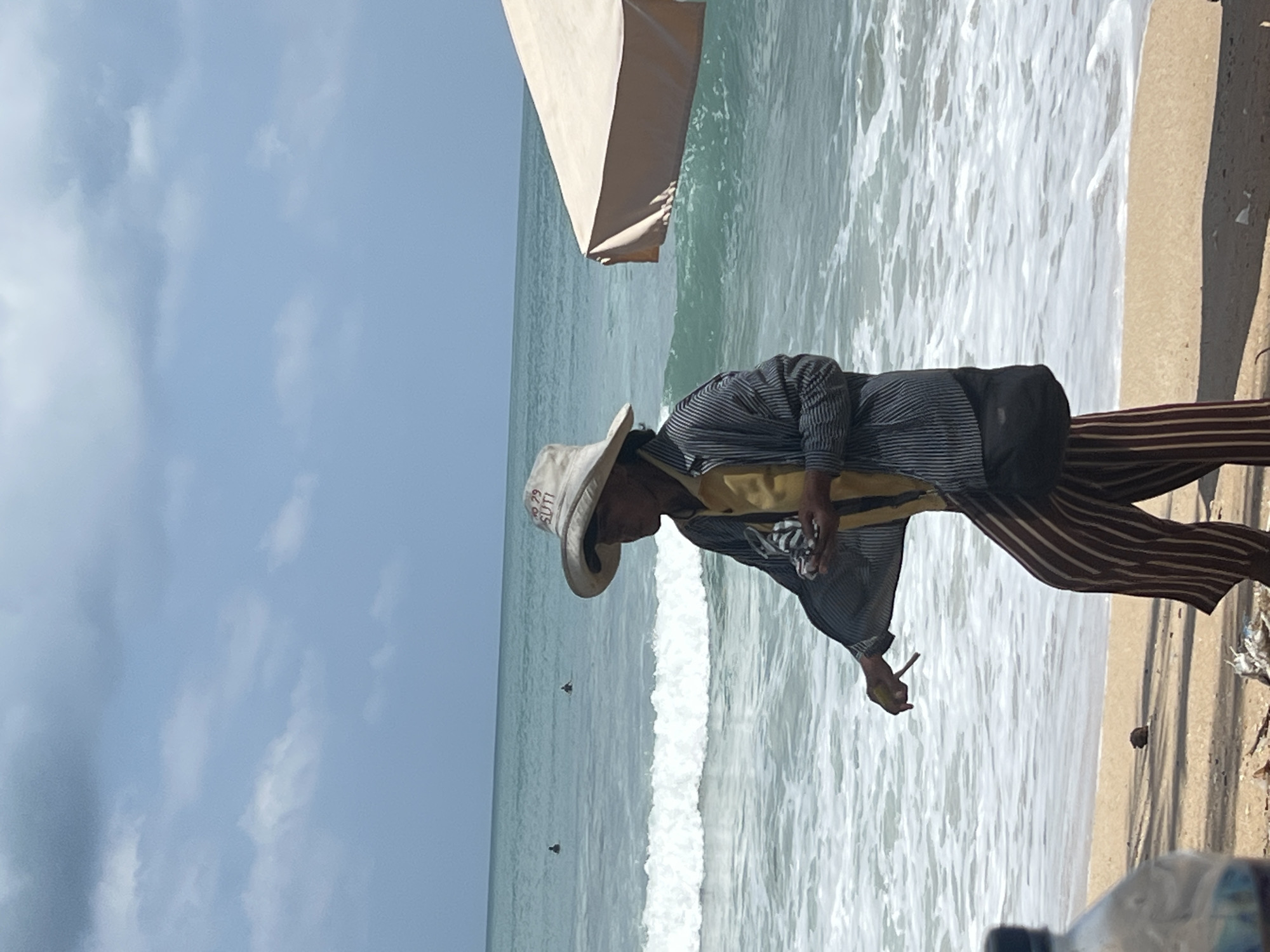 A person wearing a wide-brimmed hat, a yellow shirt, a striped jacket, and striped pants stands on a sandy beach in Kuta, Indonesia. The person is holding a small object in their hand and appears to be walking. In the background, the ocean has waves breaking on the shore, and two individuals can be seen in the water. A light-colored umbrella is visible on the right side of the image. The sky is overcast with some blue patches.