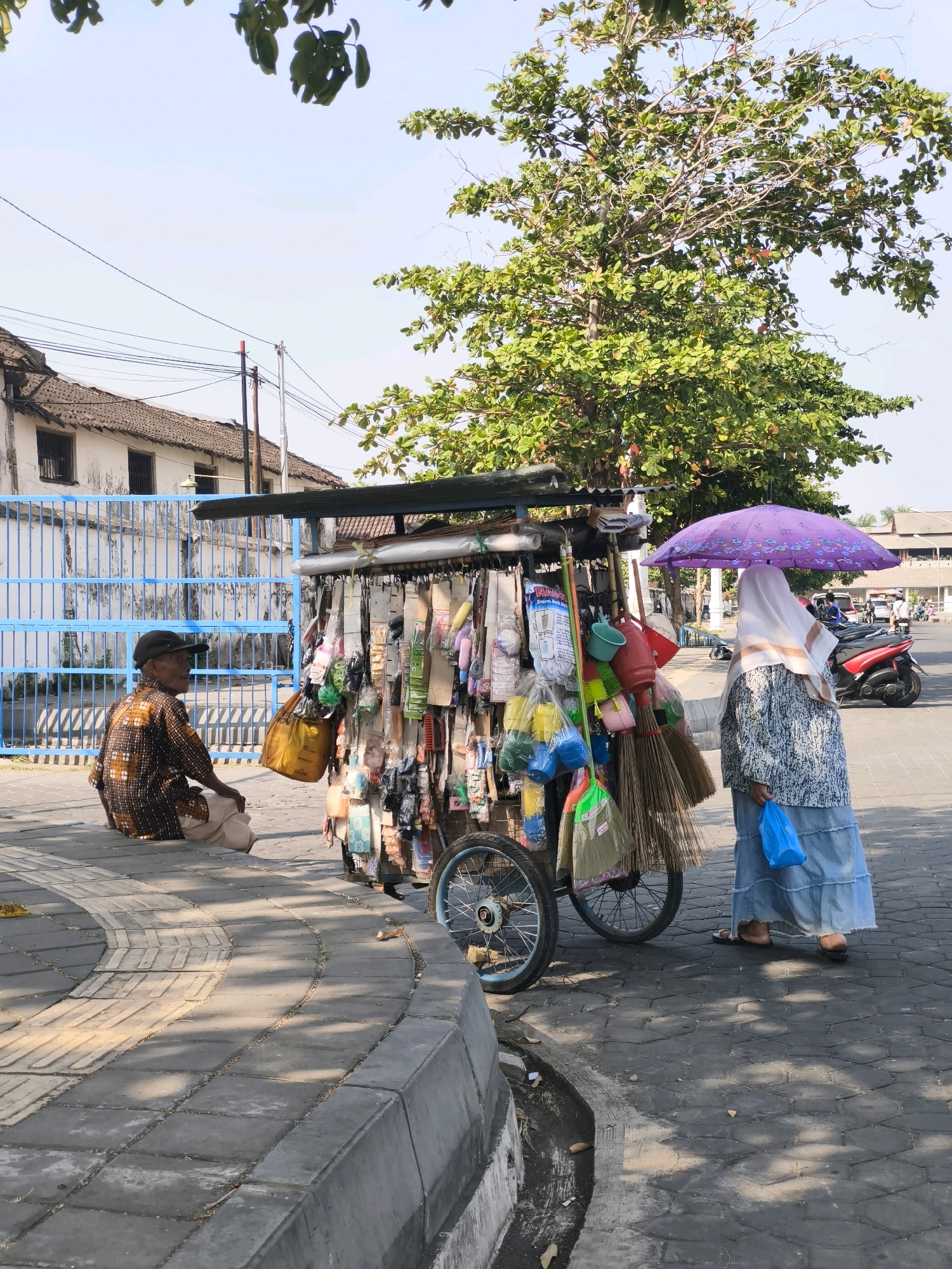 The image depicts a street scene in Surakarta, Indonesia, under a clear sky. In the foreground, a vendor's cart is laden with various household items, including brooms, brushes, and plastic bags. To the left of the cart, an elderly person wearing a patterned shirt and a cap is seated on a curb, facing away from the viewer. To the right, a woman in a headscarf and a patterned dress is standing, holding a blue plastic bag and a purple umbrella overhead. The background shows a building with a white facade and a blue fence, along with trees and a red motorcycle. The ground is paved with cobblestones, and a gutter runs along the edge of the sidewalk. The scene appears to be an everyday street market or vendor area.