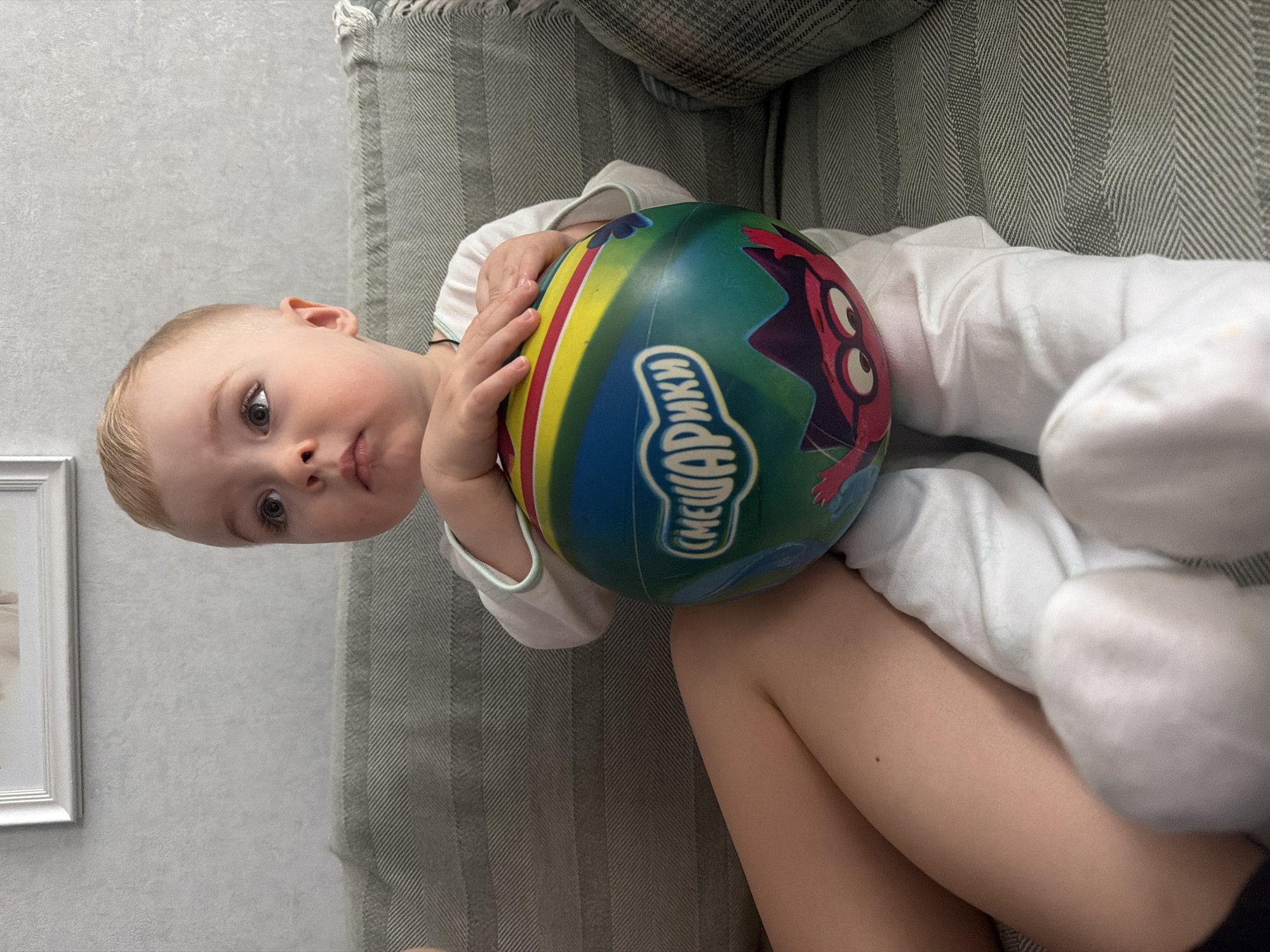 This image captures a young toddler, likely a boy, with fair skin, light blonde hair, and light-colored eyes, looking directly at the viewer with a curious, neutral expression. The child is dressed in a white long-sleeved top and white pants or socks. Due to the image's 90-degree rotation, the toddler appears to be lying on their left side.
The child is holding a brightly colored, striped ball with both hands. The ball features prominent shades of blue, green, yellow, and red, along with a cartoon character—a red creature with large, expressive eyes. Visible on the ball is Russian text, "СМЕШАРИКИ" (Smeshariki or Kikoriki), which is the title of a popular Russian animated television series for children.
The setting is indoors, with the toddler positioned on a grey upholstered sofa or couch that features a distinctive herringbone or chevron pattern. Behind the child and sofa is a light grey wall with a subtle textured finish. A white picture frame is partially visible on the wall in the lower left corner. The scene depicts a calm home environment, consistent with the provided location context of Votkinsk, Russia, especially given the Russian text on the ball. The lighting is soft and appears to be indoor, and the time of day is not discernible.