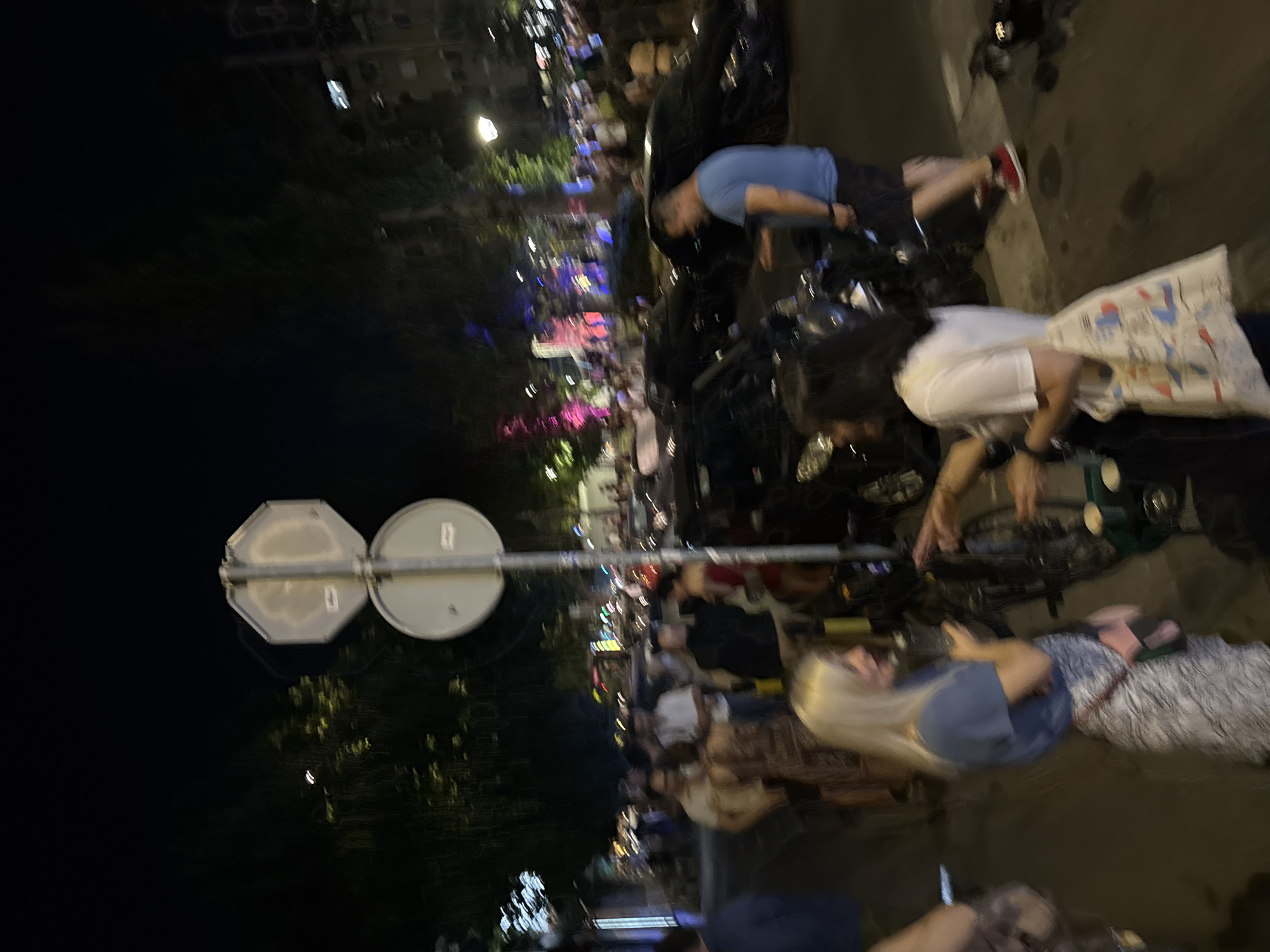 The image depicts a street scene at night, likely in Vračar Urban Municipality, Serbia, given the context provided. In the foreground, a blonde woman in a blue t-shirt and a patterned skirt is holding a drink and looking to her right. Next to her, a woman with dark hair is wearing a white t-shirt and carrying a tote bag. Behind them, several other people are visible, some standing and conversing, while others appear to be walking. Two black cars are parked on the street, one of which appears to have a damaged front end. A traffic sign with two circular signs is mounted on a pole in the middle of the scene. The background shows buildings and trees, with streetlights illuminating the area. The overall atmosphere suggests a lively evening or night out, possibly a busy street with bars or restaurants.