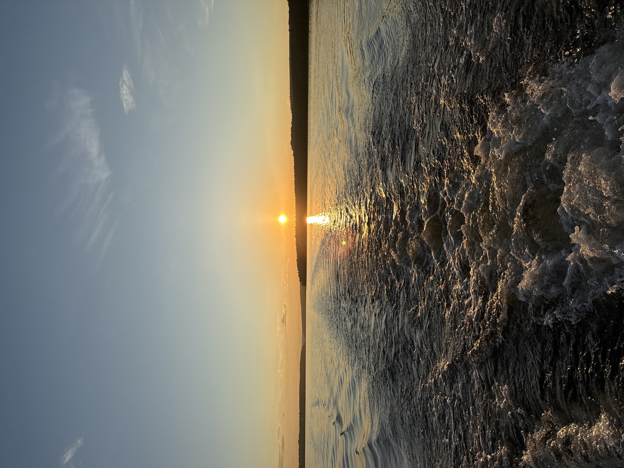This image depicts a sunset over a large body of water, likely the Votkinsk Reservoir in Votkinsk, Russia, observed from the stern of a moving boat. The sun, a brilliant orange sphere, is positioned low on the horizon towards the right side of the frame, casting a strong, golden reflection that stretches across the water directly towards the viewer. A dark, forested landmass forms a silhouette against the warm glow of the sunset along the horizon. The sky transitions from a clear, light blue at the top to a pale orange-yellow near the horizon, with sparse, wispy cirrus clouds visible in the upper sections. In the immediate foreground, the turbulent, frothy white and dark water of a boat's wake dominates, with golden highlights from the sun reflecting off the disturbed surface. Further from the wake, the water exhibits more orderly ripple patterns, also catching the ambient light. No individuals or specific man-made structures are visible within the scene, beyond the implicit presence of the boat generating the wake.