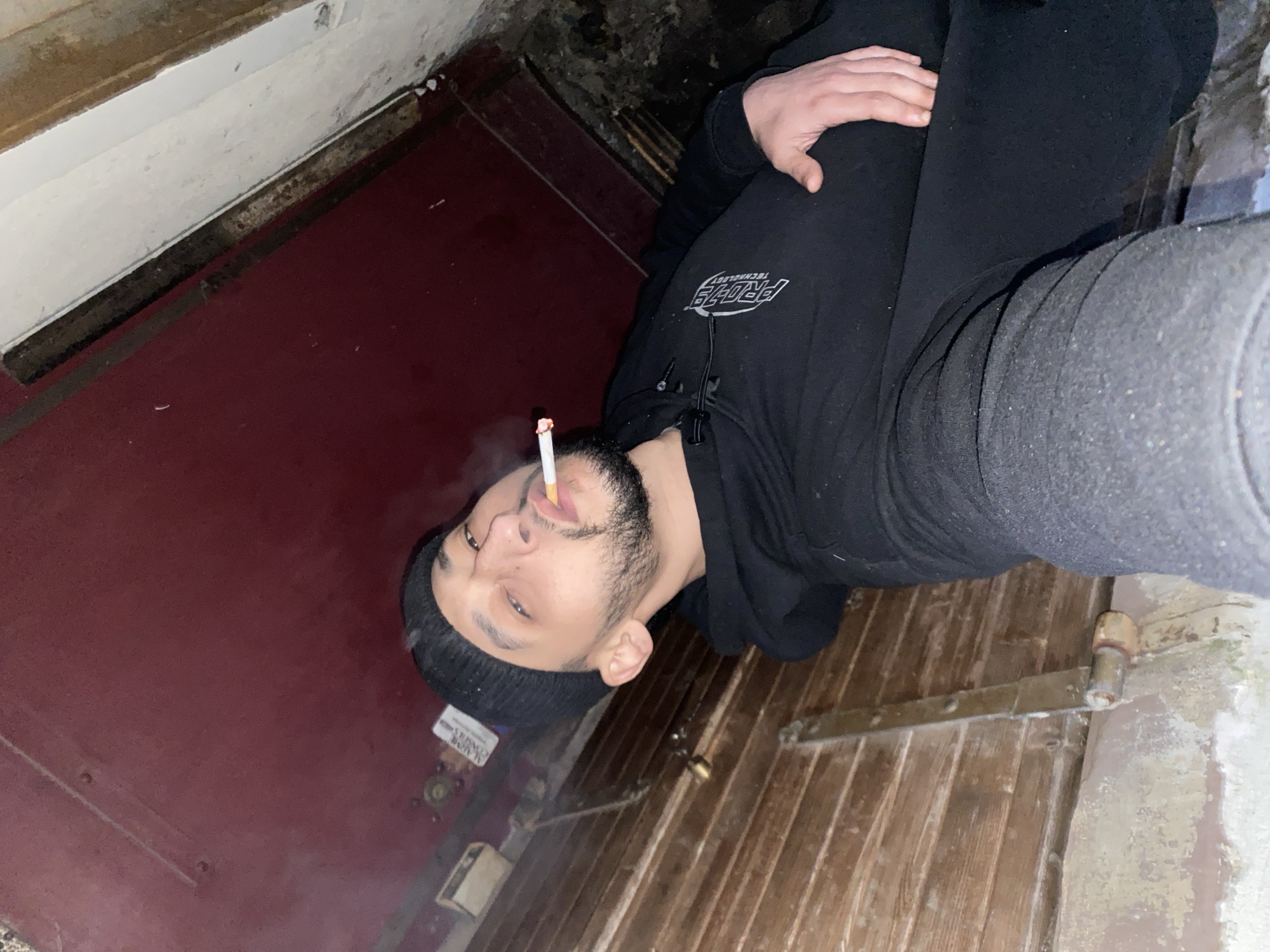 A person wearing black clothing and a black beanie is smoking a cigarette. The individual appears to be leaning against a wall or door. The location is unknown, the image does not provide enough context to identify the location. It's not possible to determine if it's in Schiltigheim, France. The background consists of a dark, possibly aged, door or wall.