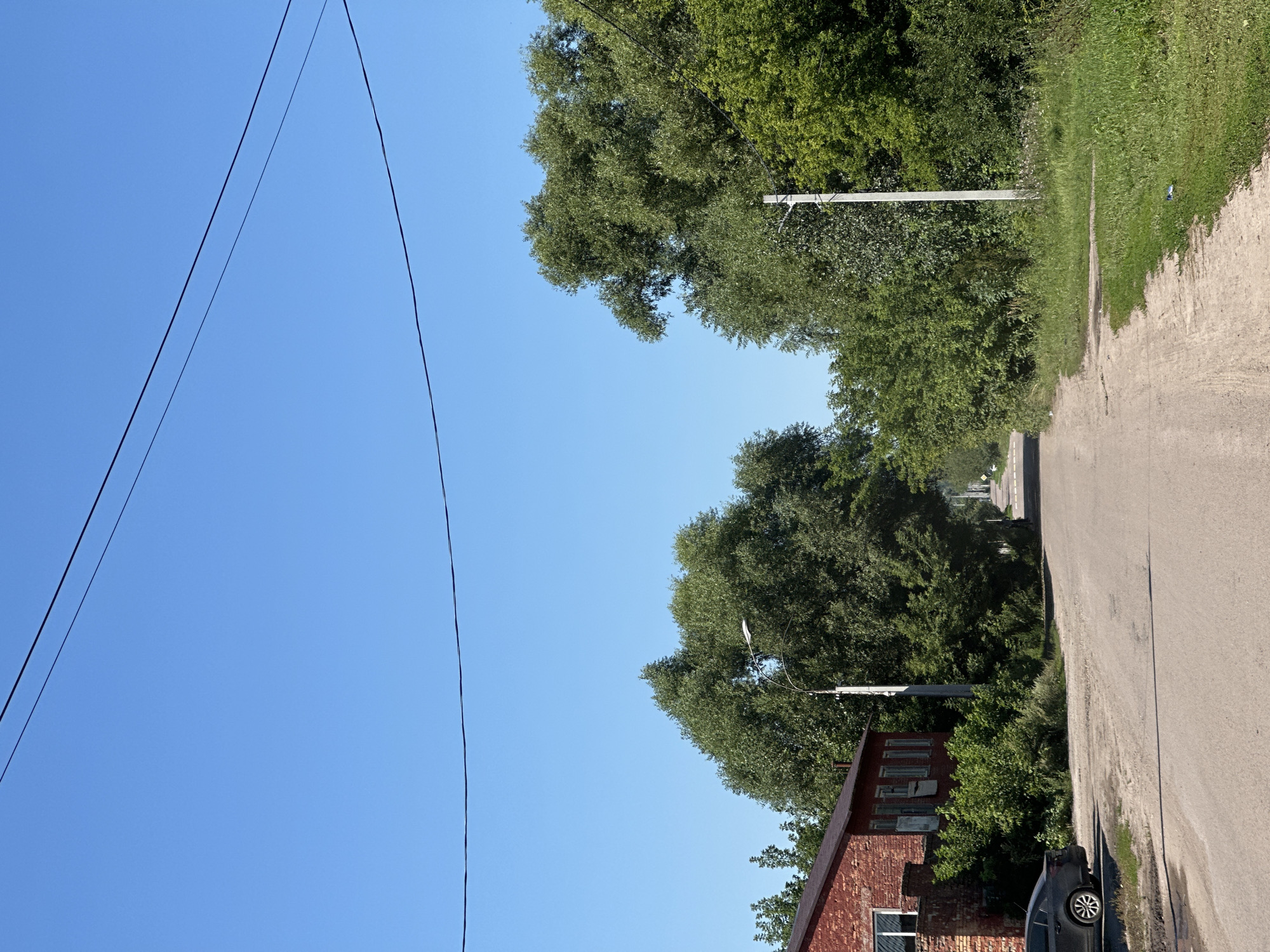 The image depicts a residential street scene in the city of Rasskazovo, Russia. A dirt road curves gently towards the background, flanked by lush green trees and vegetation on both sides. On the left, a red brick building with multiple windows and a dark roof is partially visible. A dark-colored car is parked on the left side of the road, in front of the building. A street lamp stands on a pole to the right of the road, amidst the trees. The sky above is clear and blue, with power lines crisscrossing the upper portion of the frame. The overall impression is that of a quiet, rural or suburban setting.