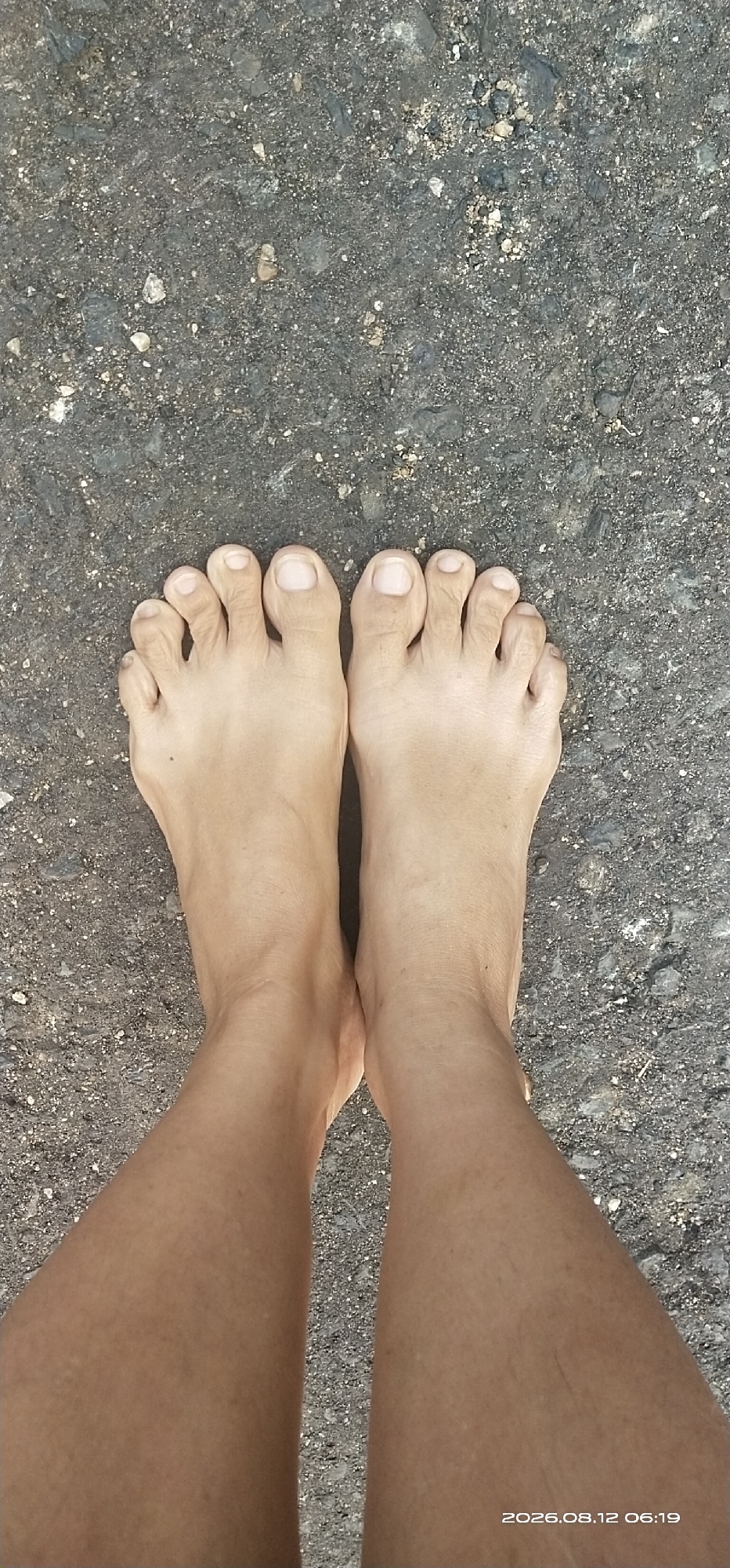 A high-angle, close-up shot shows a person's bare feet and lower legs standing on a rough, dark asphalt surface. The surface is speckled with small grey and white stones. The skin tone of the legs and feet is light brown. The toes on both feet are visible, with toenails that appear unpolished. The timestamp "2026.08.12 06:19" is overlaid on the lower right portion of the image, suggesting a specific time and date. The background, which is the asphalt, is consistent and shows no distinguishing features. The scene appears to be outdoors, possibly a street or walkway, given the asphalt ground. There is no indication of any specific location within Matabu, Indonesia, beyond what is implied by the context provided.