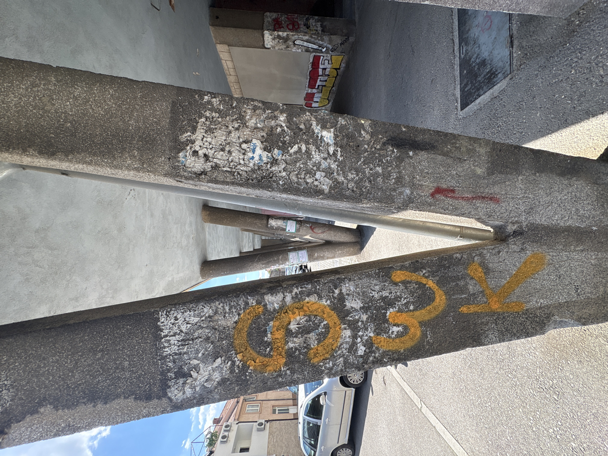 The image shows a close-up view of a concrete pillar in Mostar, Bosnia and Herzegovina. The pillar has the graffiti "003k" spray-painted in yellow. The pillar is weathered and has remnants of old stickers and posters. A narrow street and buildings can be seen in the background. A white car is parked on the street. The sky is visible in the lower left corner, suggesting it is daytime.
