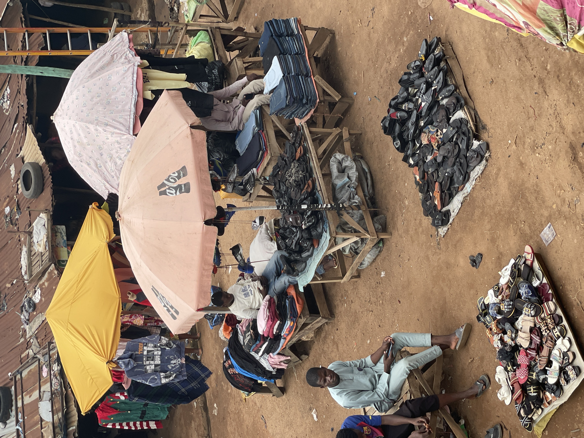 This image captures an outdoor market scene in an unknown city, in an unknown country. The ground throughout the scene is reddish-brown dirt. In the background, several structures with weathered, corrugated metal roofs are visible; one roof has a discarded tire and other debris on it. A wooden ladder leans against a building on the right side of the frame.
The market comprises multiple makeshift wooden stalls and displays. Two prominent umbrellas provide shade: a yellow one on the left and a peach-colored one with a "Coca Cola Real Taste" logo visible on the right.
Under the yellow umbrella, various clothes are displayed, including a blue hooded sweatshirt with "TIGERS" text and patterns, and a green and red striped garment.
In the midground, three men are seated on wooden benches or platforms. On the left, two men sit on a wooden bench. The man closer to the left edge wears a blue and red FC Barcelona jersey and dark shorts; only his upper body and legs are partially visible. Next to him, a man wears a light mint-green long-sleeved traditional tunic and matching pants, with grey slip-on sandals, looking to the right while holding a mobile phone.
Further to the right, under the peach umbrella, a vendor wearing a white t-shirt and blue jeans sits on a wooden platform, looking down at an object in his hands, possibly a phone. Surrounding him are stacks of folded clothing, including pink, red, blue, and black garments, displayed on a wooden table. To his right, a large pile of black shoes and sandals is arranged on another small wooden table. Behind him, partially obscured by merchandise, another vendor sits, with multiple stacks of folded denim jeans (various shades of blue) and another pile of black shoes/sandals displayed in front of him.
In the foreground on the ground, two distinct displays of footwear are present. On the left, a rectangular array of numerous colorful sandals and slippers is laid out on a white cloth. To the right, a pile of black men's shoes, including loafers and slip-ons, is arranged on a small wooden pallet.