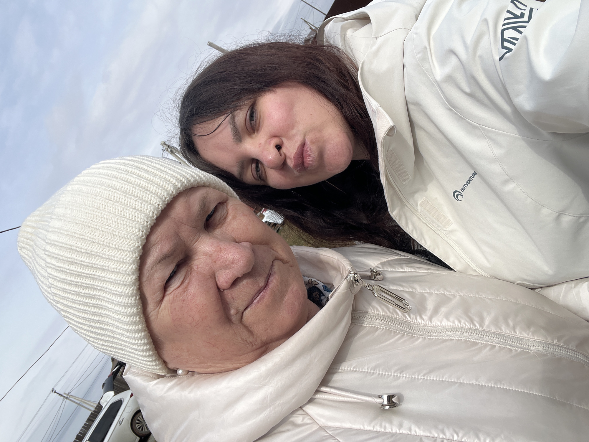This close-up selfie captures two women outdoors under an overcast sky, taken in Protasy, Russia. The image is tilted, giving a dynamic angle.
On the left, an older woman is visible from the shoulders up, wearing a cream-colored ribbed knitted beanie and a light-colored quilted jacket. She has fair skin, a gentle smile, and a small white pearl earring is visible near her ear. Her expression appears content and warm.
To her right, slightly behind and above her, a younger woman with long dark hair leans in. She is wearing a light-colored, possibly white or cream, jacket. Her face is turned towards the camera, and she is making a playful pouting expression with her lips. The younger woman's jacket features a "OUTVENTURE" logo and text prominently displayed on the front, and a darker, geometric or abstract pattern is visible on her shoulder/sleeve area.
The background, largely out of focus, indicates an outdoor setting with a cloudy sky. Faint outlines of what appears to be a white car and utility poles or power lines are visible, suggesting a semi-urban or rural environment. Both women are dressed in warm outerwear, implying cool weather. The overall mood conveyed is affectionate and lighthearted, likely a family or close friendship moment.