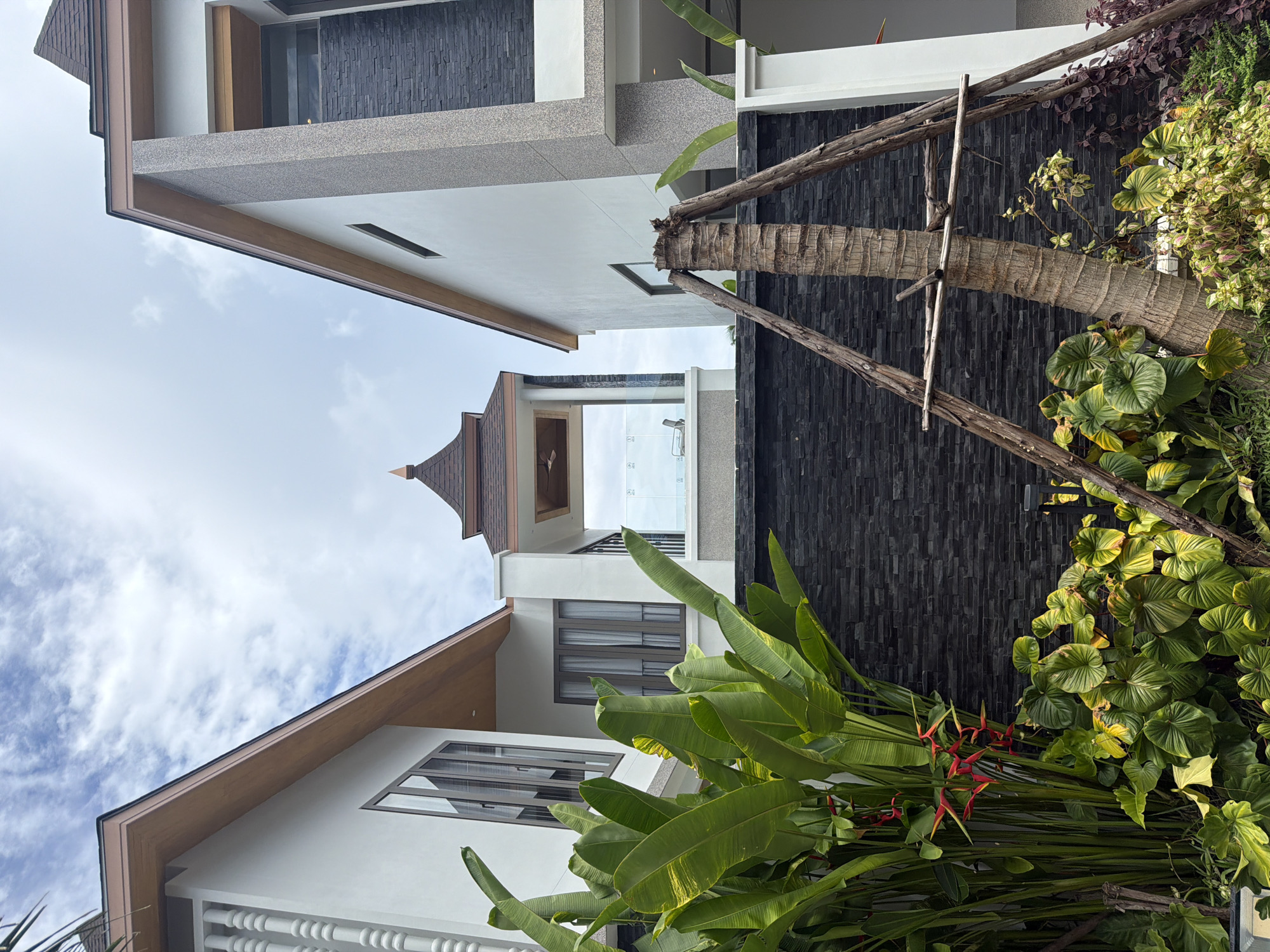 The image captures an outdoor scene in Thep Krasatti, Thailand, focusing on the architecture and landscaping of a property. In the foreground, lush tropical foliage, including large green leaves and red heliconia flowers, dominates the lower half of the frame. A palm tree trunk, secured by three wooden stakes forming a tripod, stands prominently in the center. Behind the vegetation, a wall constructed of dark, rectangular stones serves as a backdrop.
The architecture consists of two modern buildings, one on the left and one on the right, flanking a central structure. Both buildings feature white exteriors and brown-trimmed roofs. The building on the left has tall, narrow windows with reflective glass and decorative white pillars along its upper edge. The building on the right has a textured gray facade with dark stone accents and a prominent rectangular window.
In the center, a distinct structure with a multi-tiered, pagoda-like roof is visible. This structure appears to be an open-air pavilion or a balcony area, enclosed by glass railings. Through the glass, a chair and a person can be faintly discerned, suggesting an interior or semi-outdoor space. The sky above is partially cloudy with a bright, overcast atmosphere. The overall impression is one of a well-maintained, upscale property in a tropical setting.