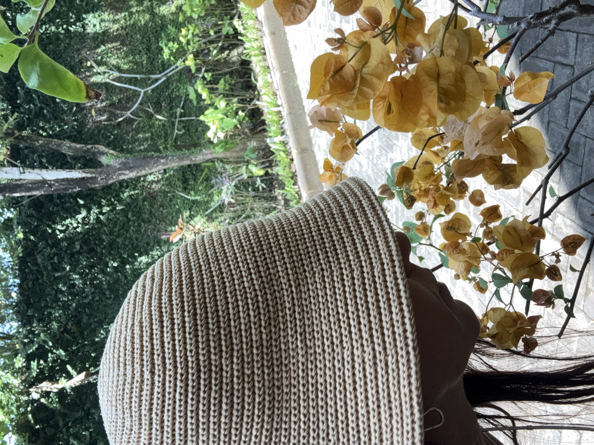 The image shows a person wearing a beige, woven hat, positioned near flowering branches with golden yellow leaves. The background includes greenery and a light-colored pavement. The setting is likely outdoors, based on the natural light and foliage, possibly within Jimbaran, Indonesia, considering the tropical vegetation and the general impression of the scene. The subject's face is partially obscured by the hat, and only dark hair is visible.