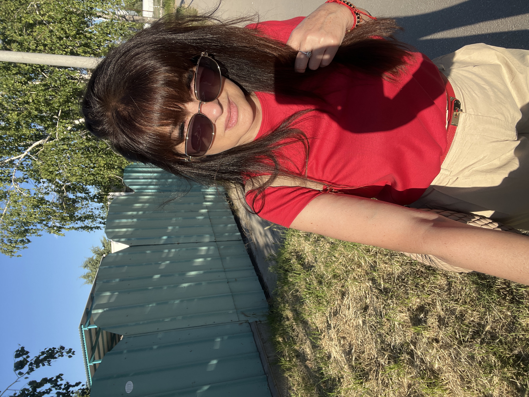 A woman with long brown hair and sunglasses poses outdoors. She wears a red shirt and beige pants with a red belt. She is holding her hair with her right hand, which has a ring on one finger and a red bracelet. The background shows a green corrugated metal structure, grass, trees, and a clear blue sky. The location is Plovdiv, Bulgaria.