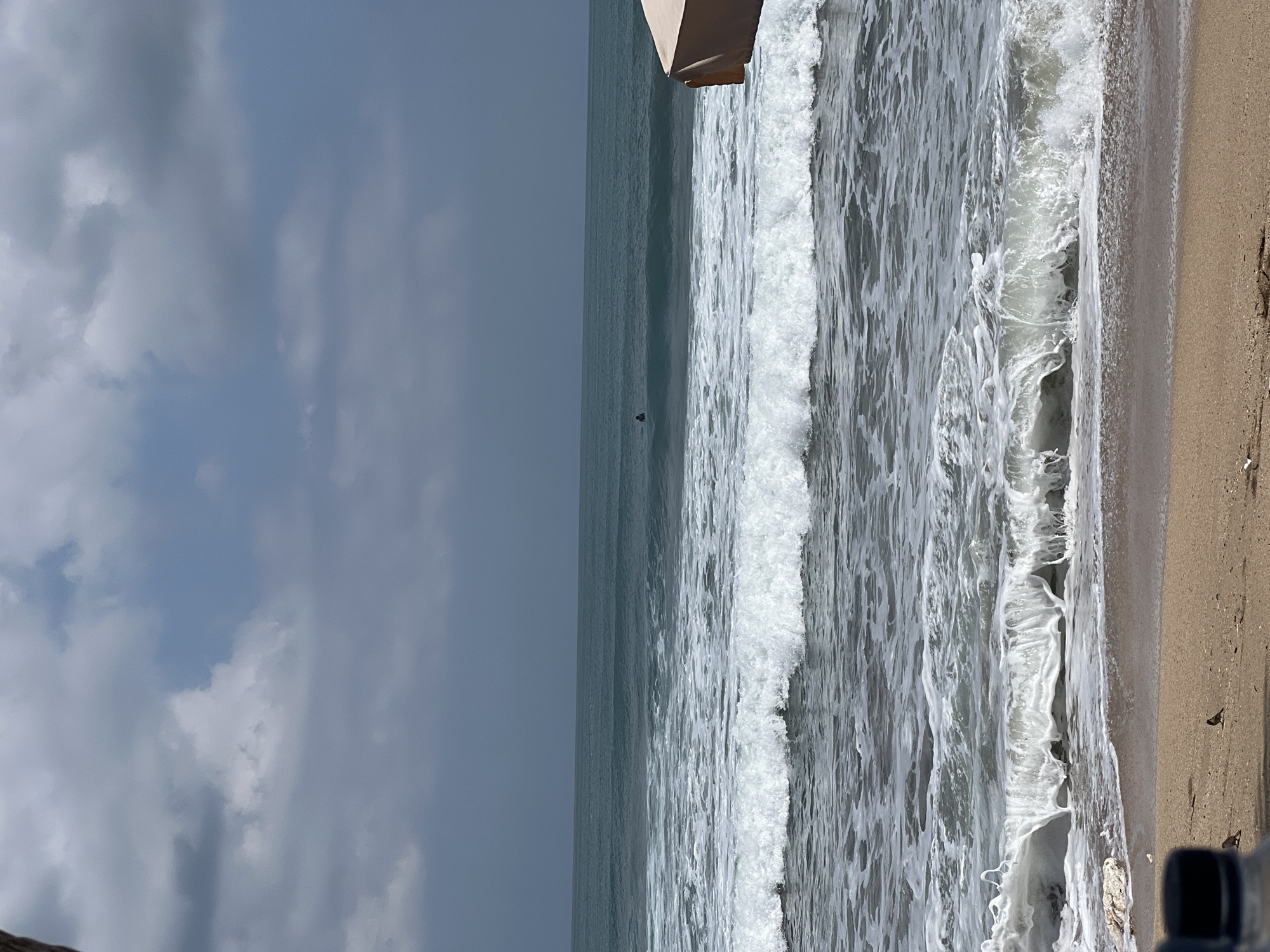 This is a medium shot, eye-level, outdoor photograph of Kuta Beach in Bali, Indonesia. The image shows the ocean with waves breaking on the shore, and a sandy beach in the foreground. The sky is overcast with some clouds visible. In the distance, there is a surfer in the water. There is a beach umbrella on the right side of the frame. The sand is light brown and there are a few small birds scattered on the beach. There is a dark blue water bottle in the bottom left corner.