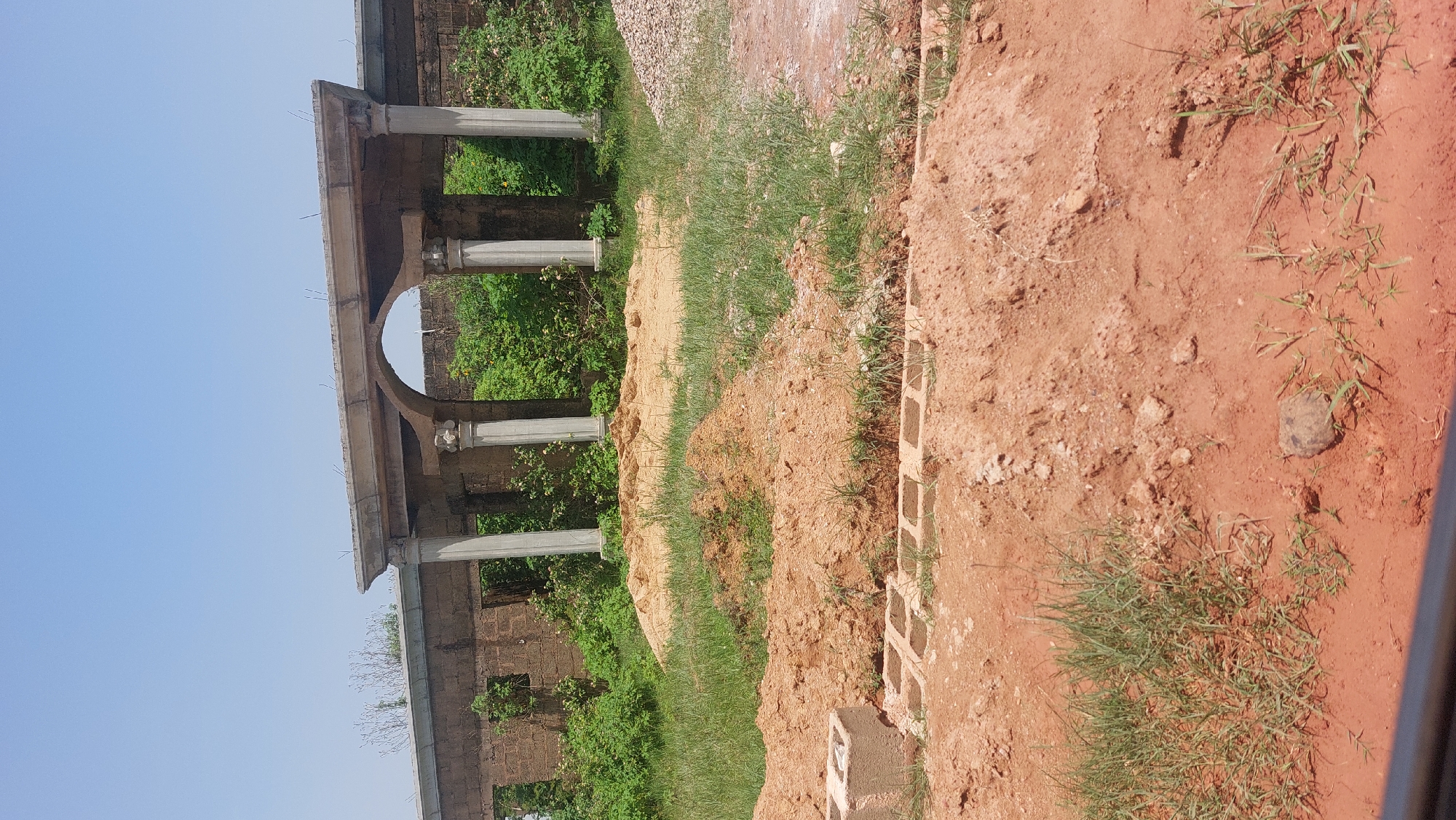 The image depicts an outdoor construction site in Gura Top, Nigeria, under a clear blue sky. In the background, a partially constructed stone wall features an arched opening supported by classical-style columns, suggesting an incomplete building or monument. Overgrown green foliage surrounds the structure and spills onto the ground. The foreground is dominated by mounds of reddish-brown soil, indicating recent excavation or material staging. Several concrete blocks are visible, some arranged in what appears to be the foundation of a new structure. A pile of grey gravel is situated on the right side of the frame. The overall scene conveys a sense of ongoing construction work in a semi-urban or rural environment.