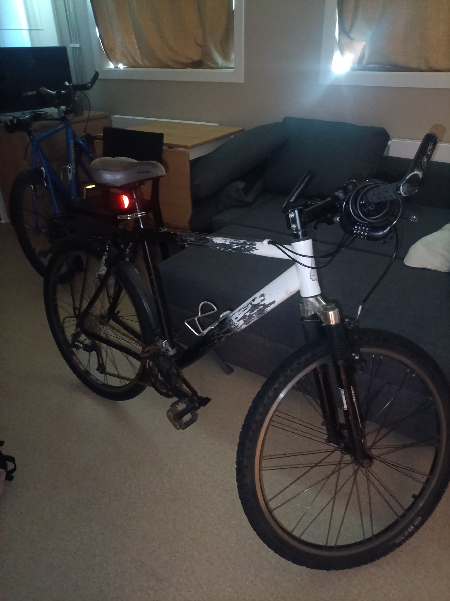 The image shows two bicycles indoors, in what appears to be a living space. One bicycle, in the foreground, is black and white with a white frame that has a distressed paint design. It has front suspension and a rear rack. A red taillight is visible on the rear of this bicycle. The second bicycle, in the background, is blue and appears to be a mountain bike. Both bicycles are parked on a carpeted floor. Behind the bicycles, a dark gray sofa is visible, with a wooden table and a window with beige curtains. The lighting suggests it might be daytime, possibly evening, with light filtering in from the window. There is no obvious activity or event taking place; the bicycles are simply parked. No people are visible in the image. There is text on the tires of the foreground bicycle and on the fork, but it is not legible. There is also a combination lock attached to the handlebars of the foreground bicycle, with numbers visible: 3335. The location context is Moss, Norway.
