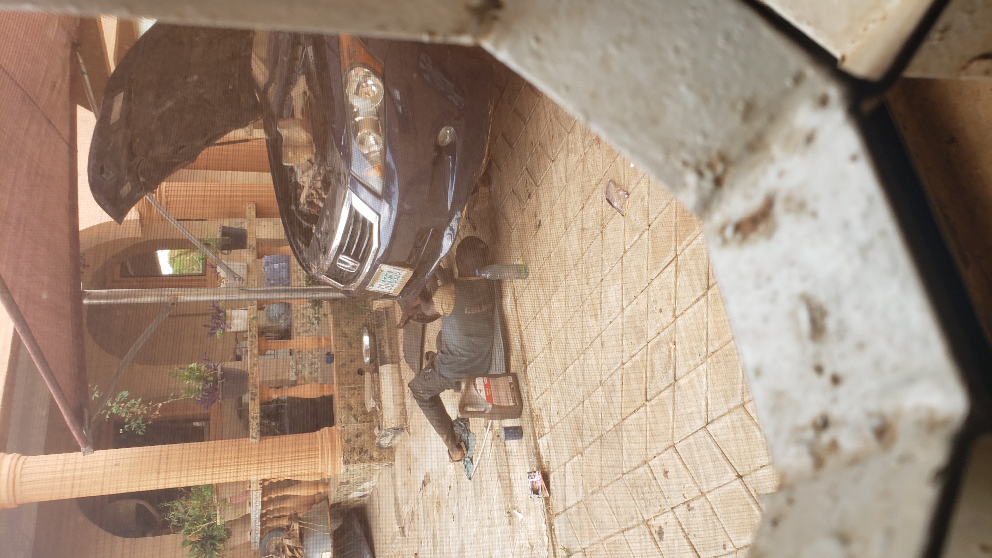 An eye-level, slightly distorted photograph shows a person working on a dark-colored car with the hood open, likely a Honda, parked in front of a house in Sabon Tasha, Nigeria. The person is dressed in casual clothing, likely doing maintenance or repairs. Scattered around the car are tools and containers, including an oil bottle. The scene is partially viewed through a window screen. Decorative columns and plants are visible in the background. The license plate of the vehicle is also visible.