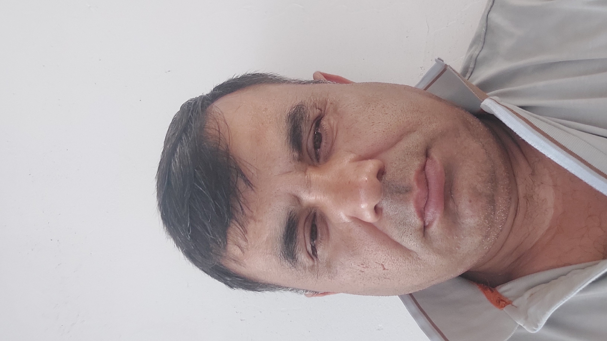 A close-up, head-and-shoulders selfie of a middle-aged man is captured against a plain white wall. The man has dark hair, dark eyebrows, and his skin appears to be tanned. He is wearing a light-colored collared shirt with striped trim. The lighting suggests an indoor environment, possibly with natural light. The scene does not contain any elements that would allow for the identification of a specific location within the city of Eski-Alimkent, Uzbekistan.