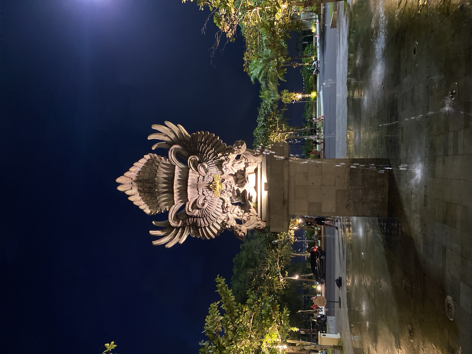 The image depicts a large stone sculpture of a mythical bird, likely a Garuda, at night in the city of Kuta Selatan, Indonesia. The sculpture is positioned in the center of a plaza with a water fountain at its base. The sculpture is illuminated from below, highlighting its intricate details and imposing wingspan. Several trees, some with string lights, line the plaza, and a street with a few cars and pedestrians is visible in the background. The sky is dark blue, indicating nighttime. The ground of the plaza is paved with stone tiles, and the water from the fountain creates reflections and splashes on the wet surface. A few people are walking and standing around the plaza, and a motorcycle is parked in the distance. The scene suggests a public space in Kuta Selatan, Indonesia, possibly a tourist area or a significant landmark, with a blend of cultural art and modern amenities.