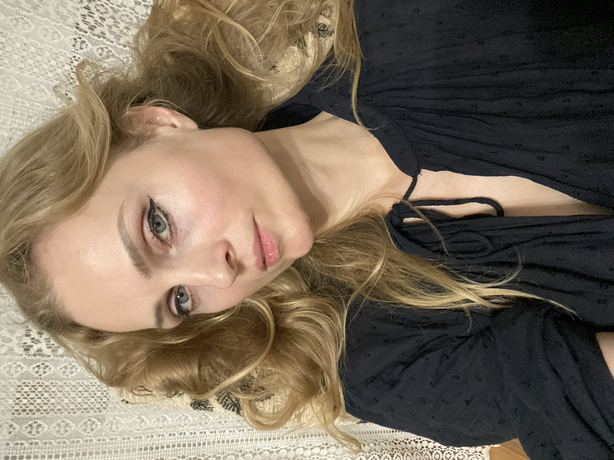 A medium shot captures a fair-skinned woman with blonde, wavy hair, wearing a dark fabric shirt. She is positioned in a reclined pose against a backdrop of white lace. Her head is turned to the side, and her gaze is directed downwards. The woman's makeup includes eyeliner and mascara, and her lips are subtly colored. The image is well-lit, with the light source appearing to be from the front. The overall tone is serene and intimate. The background suggests an indoor setting, possibly a home, with the lace providing a decorative element. There are no clear indicators of the specific location within the city of Lida, Belarus.