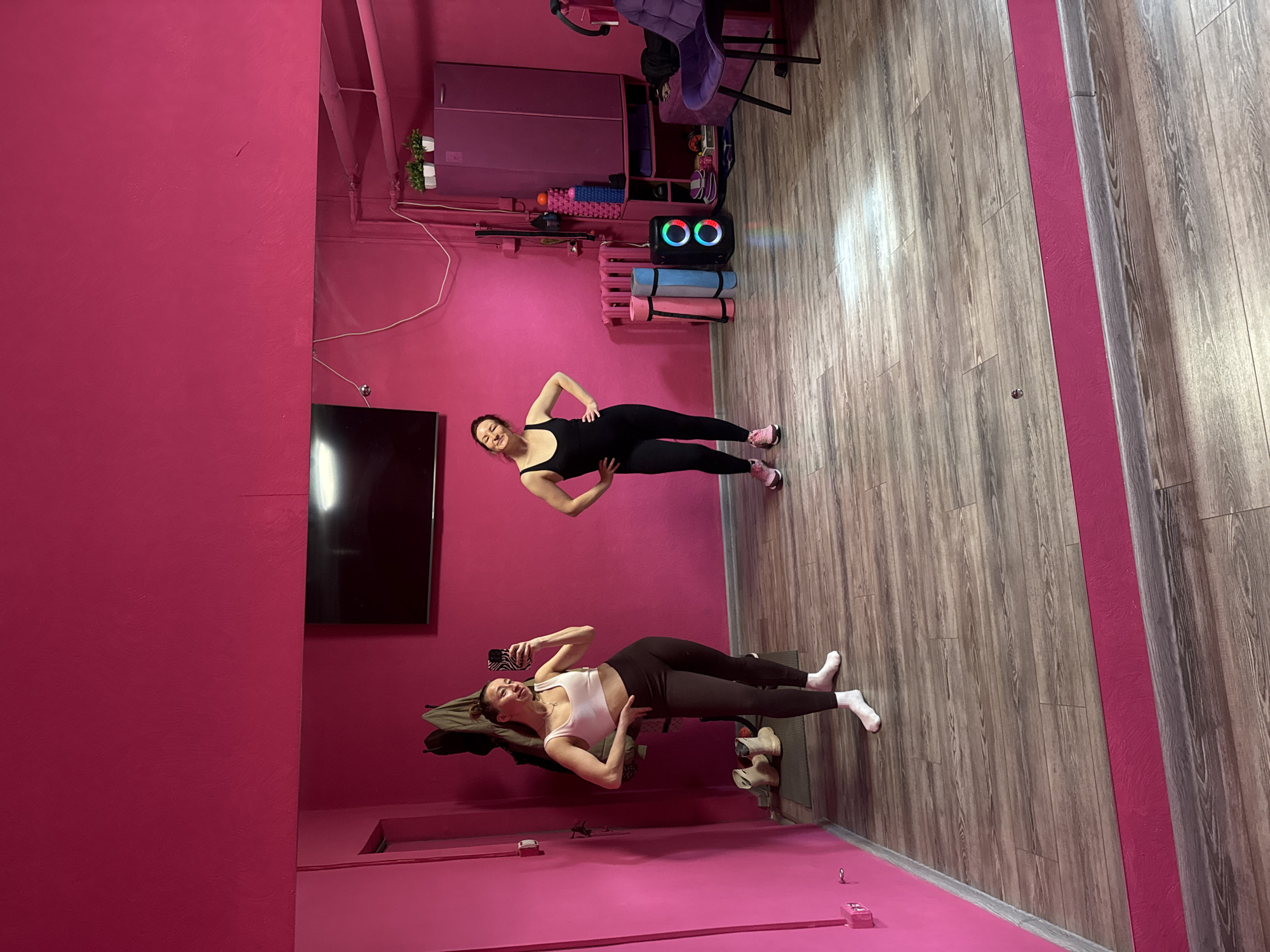 Two women are in a room in Chaikovsky, Russia. The room has pink walls and a wood-patterned floor. One woman, wearing a white top and dark leggings, is lying on her side and taking a selfie with her phone. She is propped up on a bench with shoes nearby. The other woman, wearing a black bodysuit and pink sneakers, is standing and posing with her hands on her hips. Behind them are a TV, cabinet, exercise equipment, and a stroller.