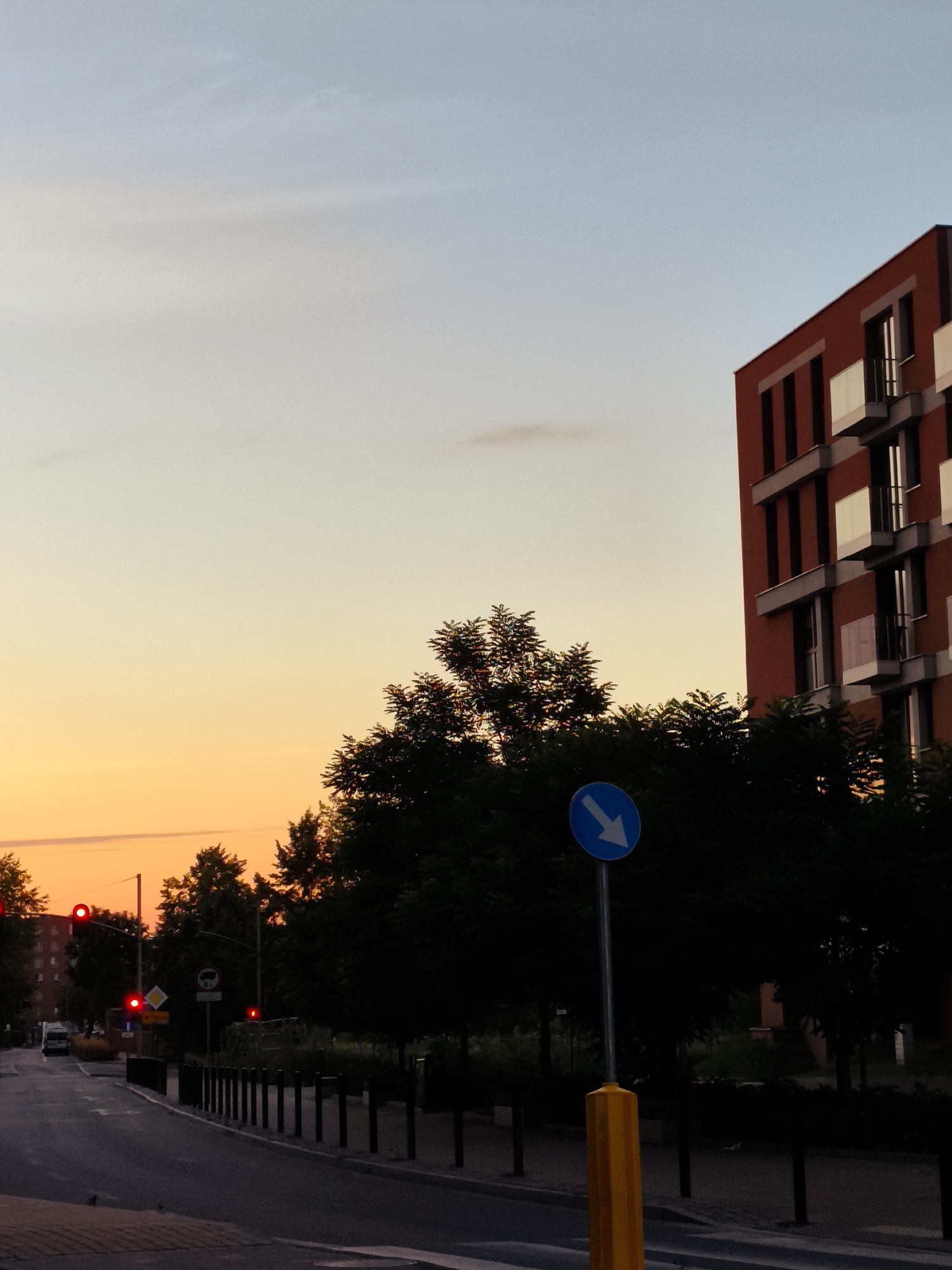 The image shows a street scene at dusk in Gdańsk, Poland. The sky is a gradient of pale blue at the top to warm orange and yellow near the horizon. On the right side of the image is a modern red-brick building with multiple balconies. In the foreground, a street runs diagonally from the bottom left towards the middle. Trees line the street on the left and in the middle ground, their dark foliage silhouetted against the sky. A blue circular road sign with a white downward-pointing arrow is visible on a pole in the middle ground. Several traffic lights with red lights illuminated are visible in the distance on the left side of the street. A few cars are present on the road. The scene appears to be an urban environment.
