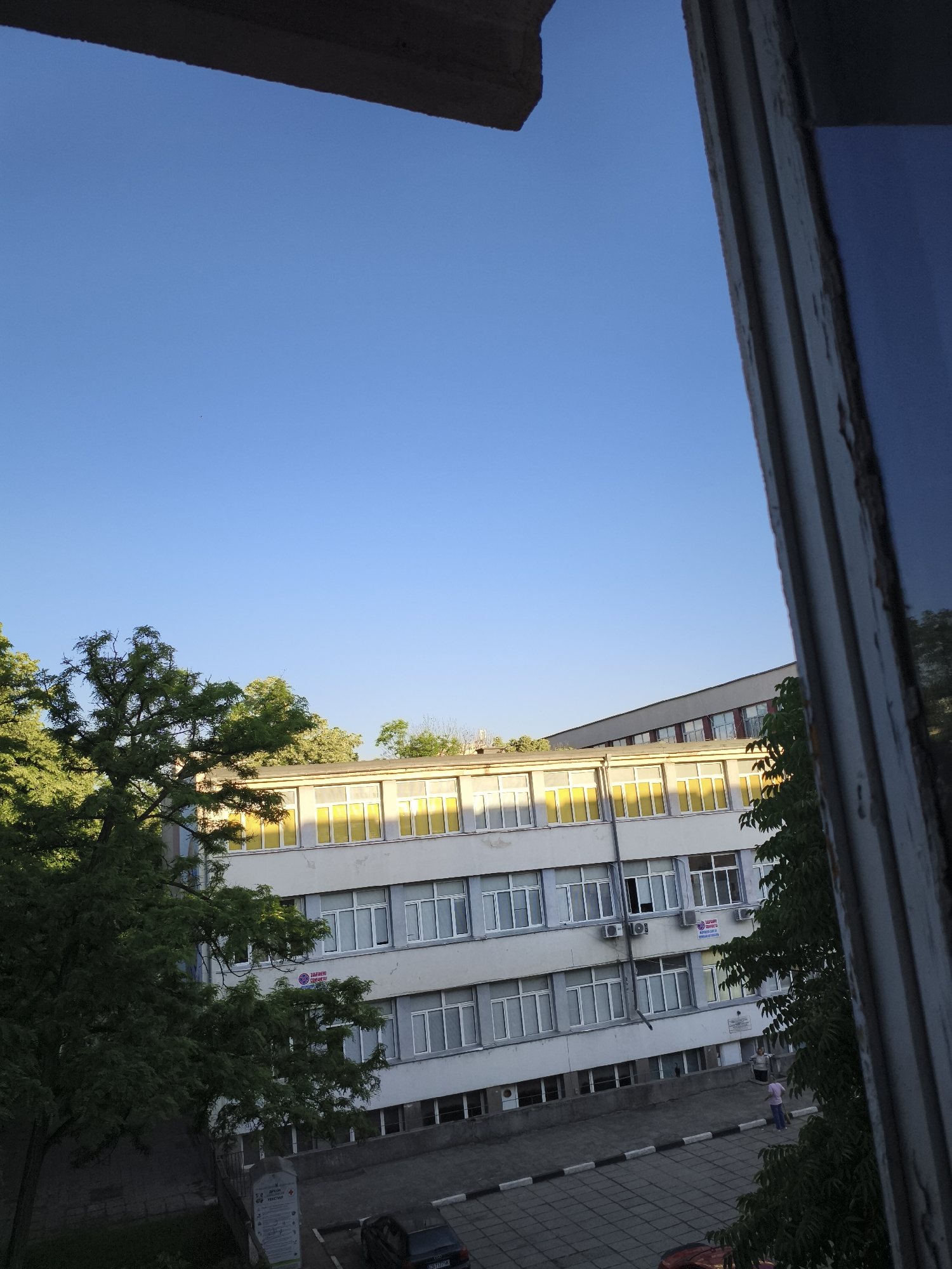 The image, taken from an elevated position through a window frame, shows a multi-story building in Plovdiv, Bulgaria, under a clear blue sky. The building is white with multiple rows of windows, some having a yellow hue suggesting interior lighting or blinds. Trees partially obscure the left side of the building. The ground area in front of the building is paved, with visible cars and a sign. Two people are visible near the building's entrance.