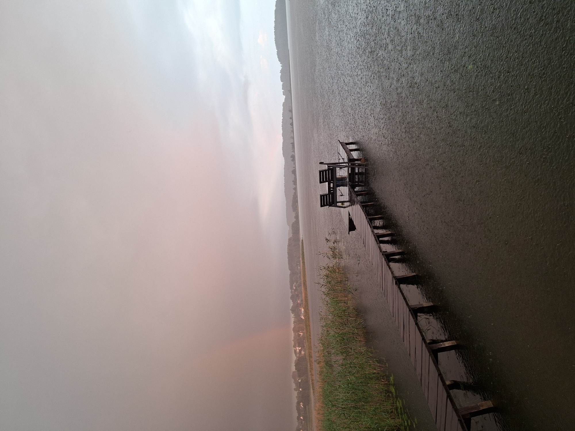 The image depicts a wooden pier extending into a dark, rain-swept lake in the Слабодкаўскі сельскі Савет, Belarus. The sky is overcast with a hint of pink and a faint rainbow is visible in the distance on the left side, suggesting a recent or ongoing rain shower. Reeds line the edge of the lake on the left, bordering the pier. At the end of the pier, two empty wooden chairs and a small table are positioned. The lake surface is disturbed by numerous raindrops, creating ripples. The far shore is lined with a dense forest and a cluster of buildings in the distance. The overall atmosphere is serene and melancholic, with the rain emphasizing the isolation of the scene.