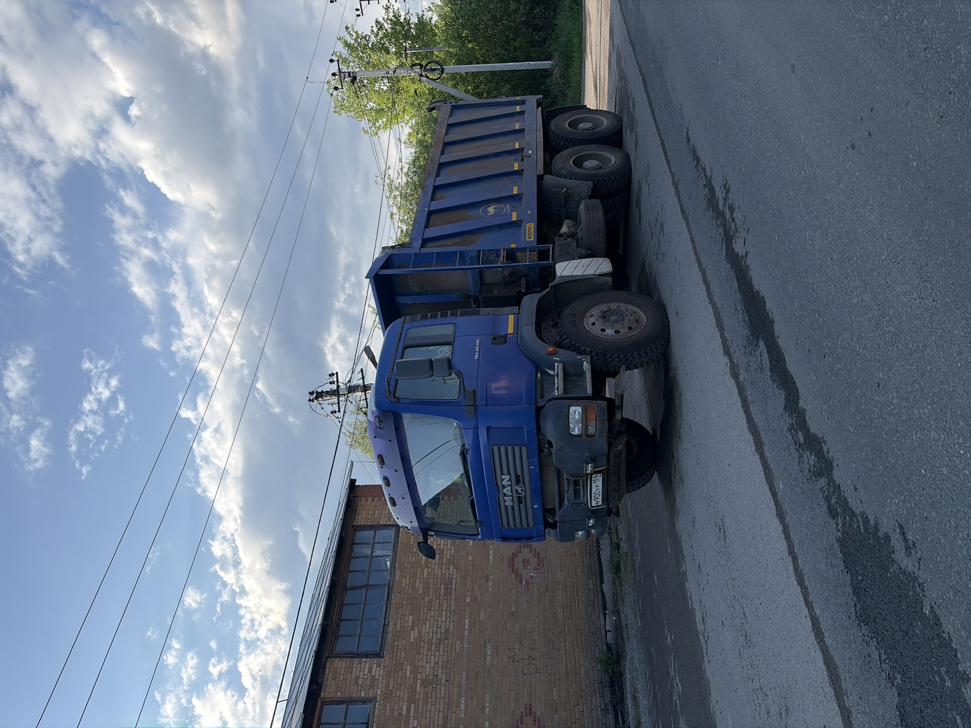 The image shows a blue MAN TGA 6x4-68.40 dump truck parked on an asphalt road next to a brick building. The truck has a license plate that reads "H900YP 159", indicating it is registered in the Perm region of Russia. The building appears to be an older industrial or commercial structure with multiple windows. Overhead, there are power lines and utility poles. The sky is partly cloudy. The environment suggests an urban or industrial area of Perm, Russia.