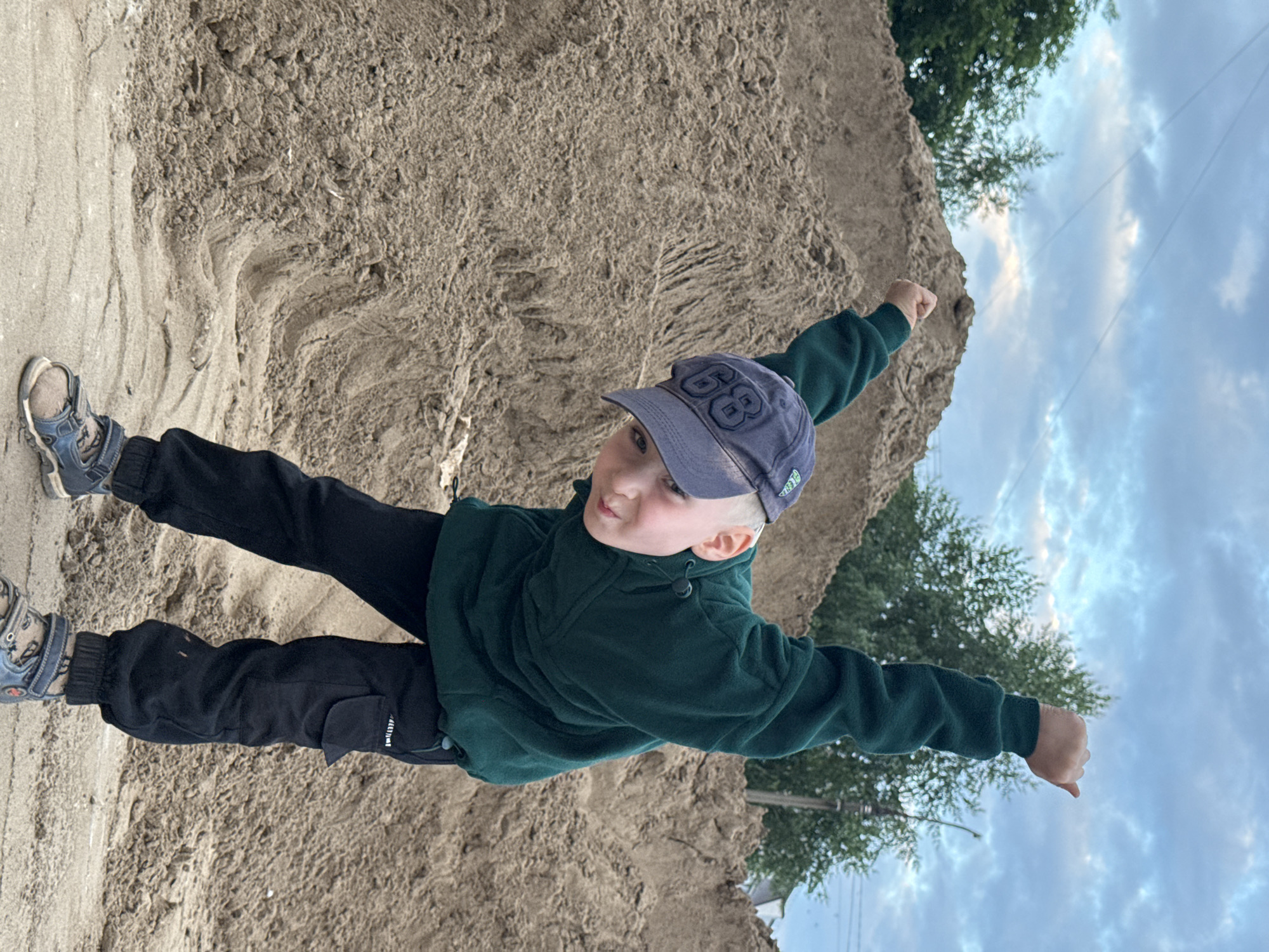 A young boy wearing a dark green hooded jacket, black pants, and a blue baseball cap with the number "68" on it, stands in a sandy area. He is facing forward, with his arms outstretched to the sides as if he is flying. His left leg is bent at the knee, and his right leg is extended behind him. The boy appears to be joyful and energetic, with a slight smile on his face. He is wearing blue and gray sneakers.
The background consists of a large mound of sand, possibly a construction site or a playground. In the upper right corner of the image, there are green trees and a cloudy blue sky, suggesting it is daytime and potentially late afternoon. The overall impression is a candid shot of a child having fun outdoors in Chaikovsky, Russia.
There is text visible on the boy's pants, appearing to be a brand name or logo, but it is difficult to read clearly. The text on his cap, "68", is prominently displayed. The image is shot from a low angle, emphasizing the boy's playful pose against the backdrop of the sand and sky.