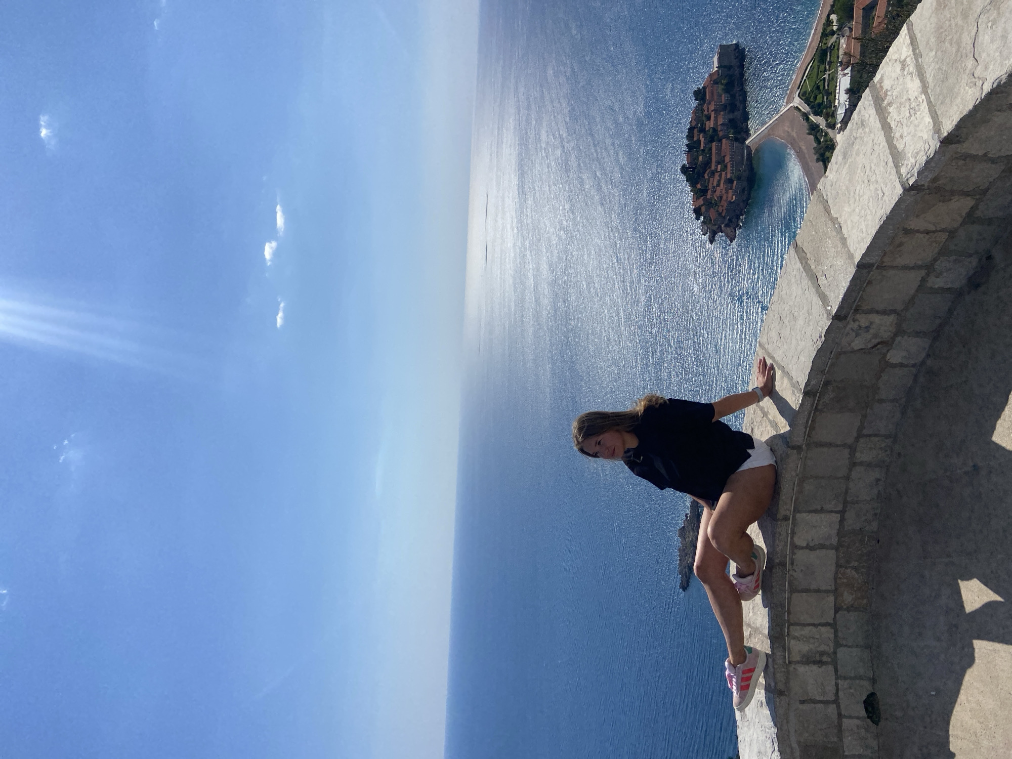 This image presents a stunning, sunlit panoramic view of Sveti Stefan, Montenegro, from a high vantage point. A young woman is the central subject, positioned in the foreground, casually perched on a low, curved stone wall or parapet. She has long, light brown hair and is dressed in a black short-sleeved top, white shorts, and pink and white sneakers, identifiable by their three stripes as likely Adidas. Her body is angled slightly, with her gaze directed towards the camera, a subtle smile on her face, conveying a sense of relaxed enjoyment.
Behind the woman, the vast, sparkling blue waters of the Adriatic Sea dominate the scene, extending to a hazy horizon line. The sky above is a brilliant, clear blue with a few faint, wispy clouds, indicating bright, sunny weather. The strong daylight casts distinct shadows on the stone wall where the woman is sitting, suggesting it's likely midday or early afternoon.
In the mid-ground, to the right of the frame, lies the distinctive fortified island of Sveti Stefan. It is densely covered with numerous terracotta-roofed buildings and some greenery, connected to the mainland by a narrow causeway. A small sandy beach is visible next to the causeway, meeting the calm, shimmering sea. The overall atmosphere is serene and picturesque, highlighting the beauty of the coastal landscape. No visible text is clearly discernible beyond the brand-characteristic stripes on the sneakers.