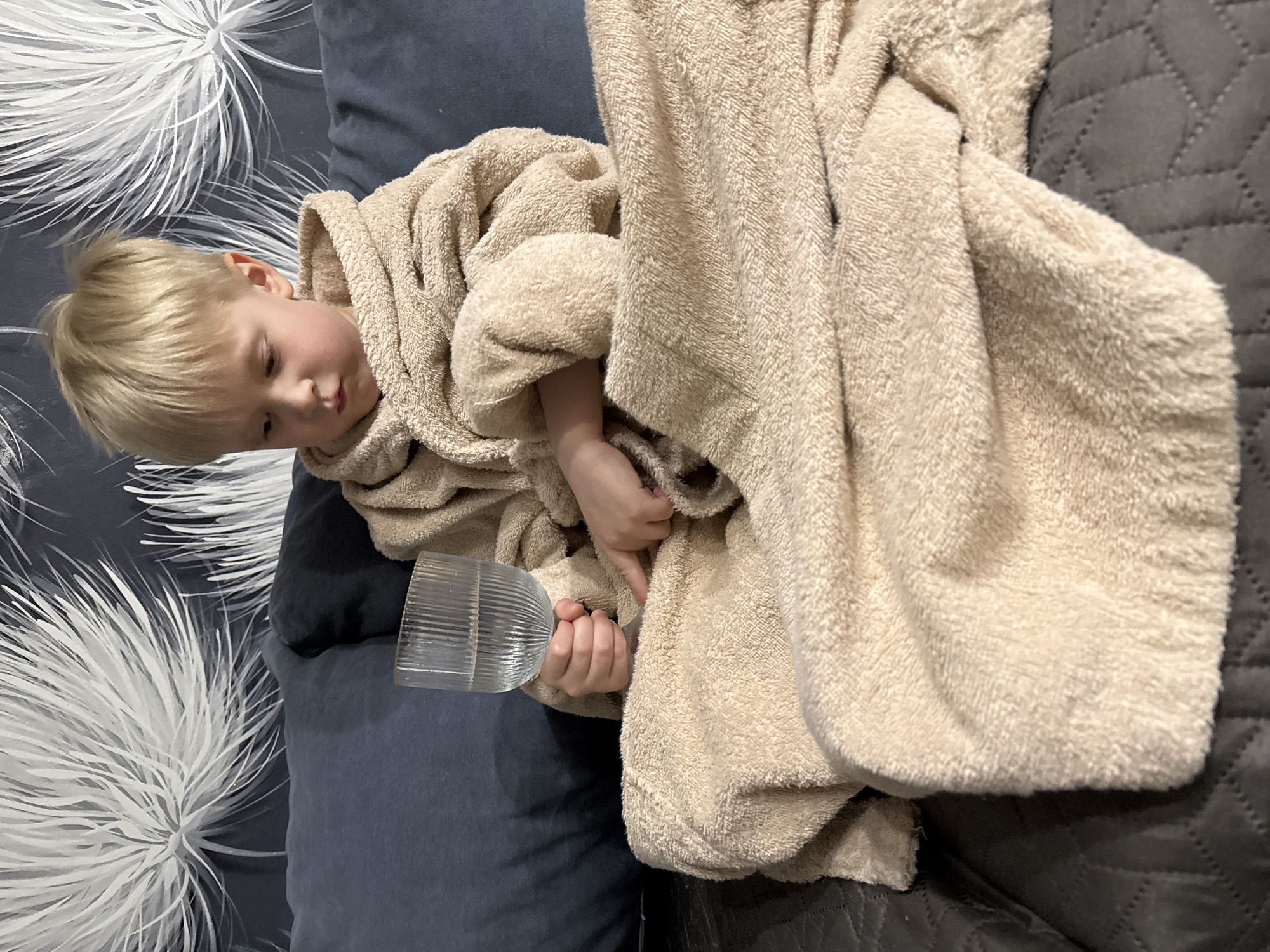 A young child with short, light blonde hair is depicted lounging on a piece of furniture, likely a couch or bed, indoors in Chaikovsky, Russia. The child is wearing a plush, light beige or cream-colored hooded bathrobe, which is draped around them, partially covering their body. They appear to be resting or relaxing, with their head slightly tilted and eyes looking down towards an object they are holding.
In the child's left hand, they hold a clear, cylindrical glass with a ribbed or fluted texture, suggesting it might contain a drink, though the contents are not visible. The child's expression is calm and perhaps a little contemplative or sleepy.
The background consists of several distinct textures and patterns. To the left and behind the child's head, there's a dark gray or dark blue surface, possibly a pillow or cushion, against which a patterned fabric is visible. This fabric features prominent white, feathery, or dandelion-like floral patterns on a dark background. To the right of the child and the bathrobe, there is another dark gray surface with a distinct quilted, geometric pattern, likely another blanket or part of the furniture. The overall lighting suggests an indoor setting, without specific indications of the time of day. No text is visible in the image.