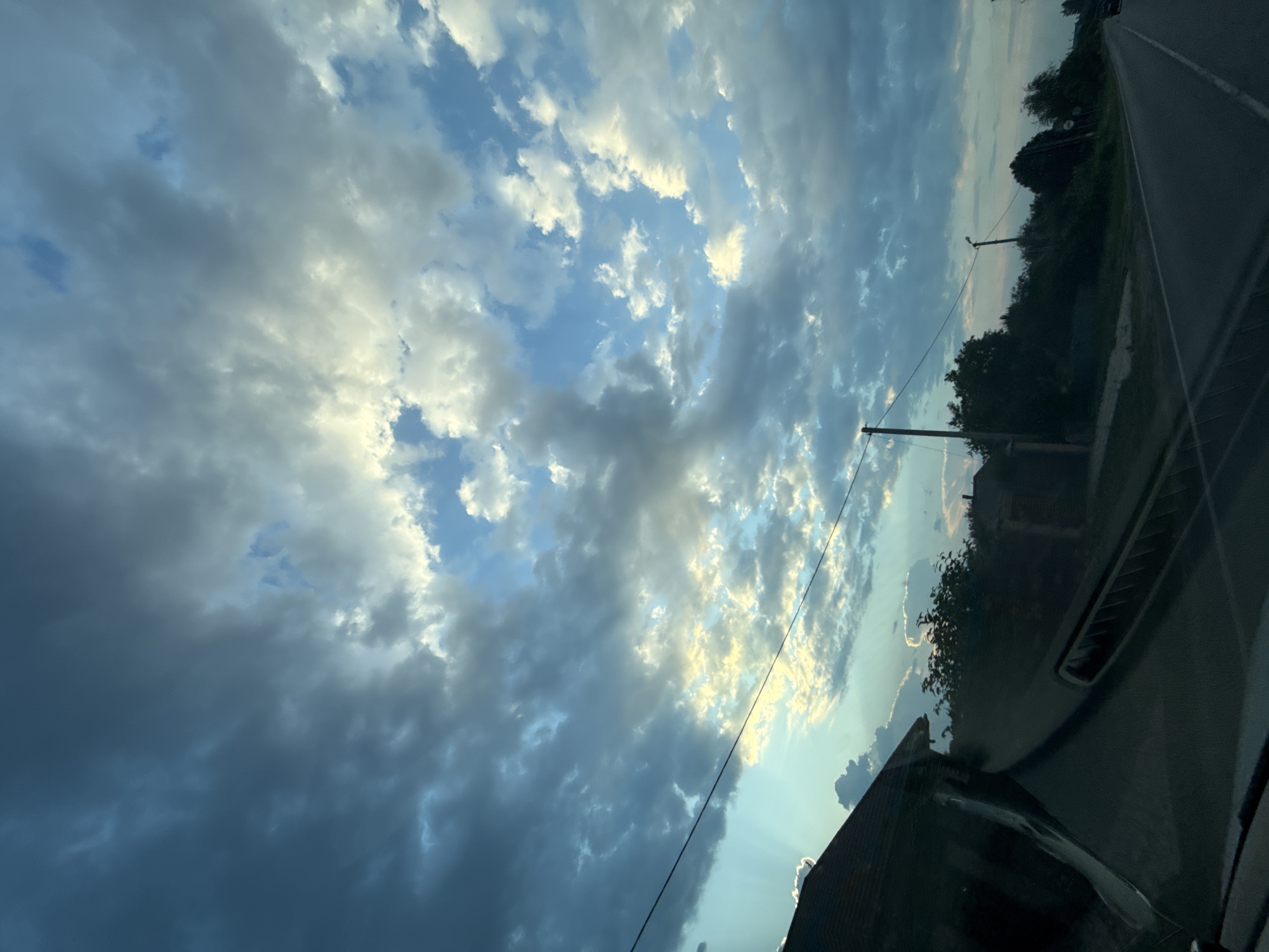 The image shows a view from inside a vehicle looking out at a rural landscape under a dramatic, cloudy sky. The sky is filled with a mix of dark, heavy storm clouds and brighter, illuminated clouds, suggesting either an approaching storm or a sunset/sunrise breaking through. Rays of light are visible filtering through the clouds on the left side of the image.
In the foreground, the dashboard of the vehicle is visible, with a dark, reflective surface. On the left, the corner of a building with a dark roof is visible, partially obscured by the vehicle's interior.
Beyond the vehicle, the landscape consists of a road stretching into the distance, with a tree line bordering its right side. Several power poles and lines are present along the road. A few buildings and structures are visible nestled among the trees. The overall environment appears to be a village or rural area, consistent with the location of Nizhneatamanskoe, Russia. No people are visible in the image.